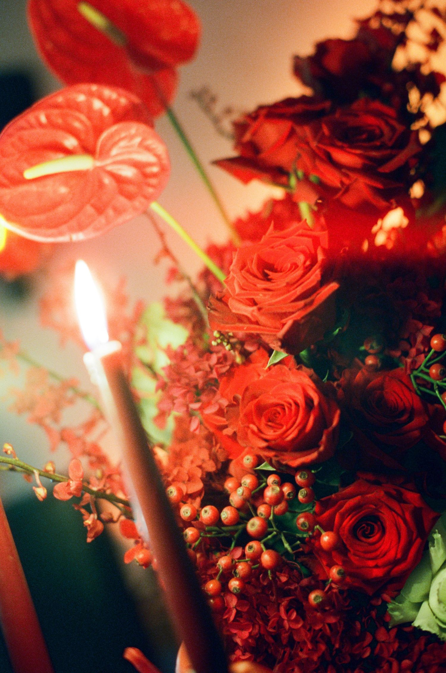 Red roses, anthuriums, and berries arranged with a lit candle in a lush floral display.