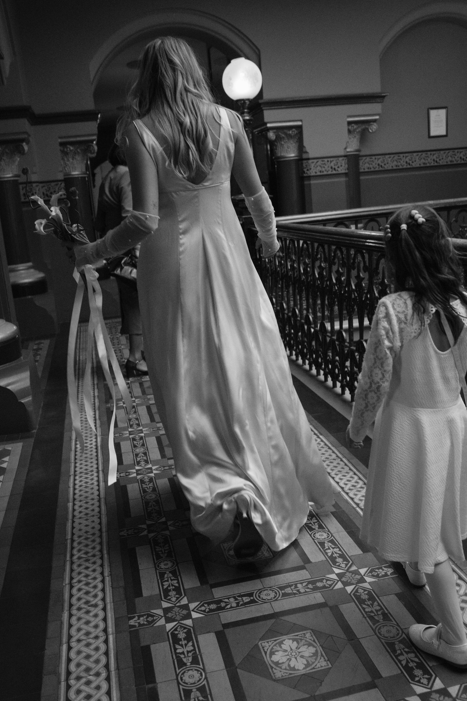 Black and white photo of a bride in a long white gown walking up ornate stairs with a young flower girl.