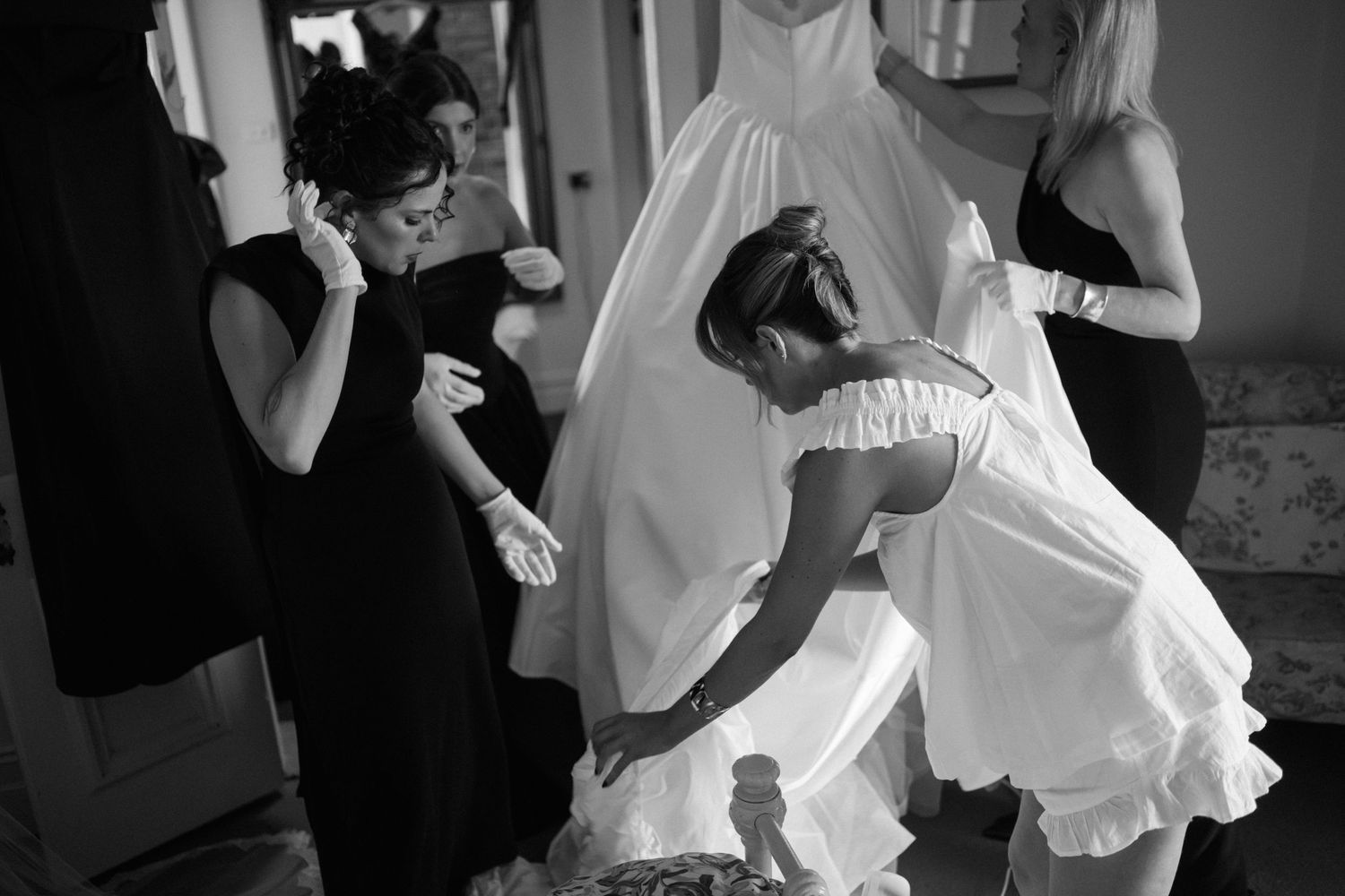 Black and white photo of bridesmaids helping adjust a wedding dress during preparations.