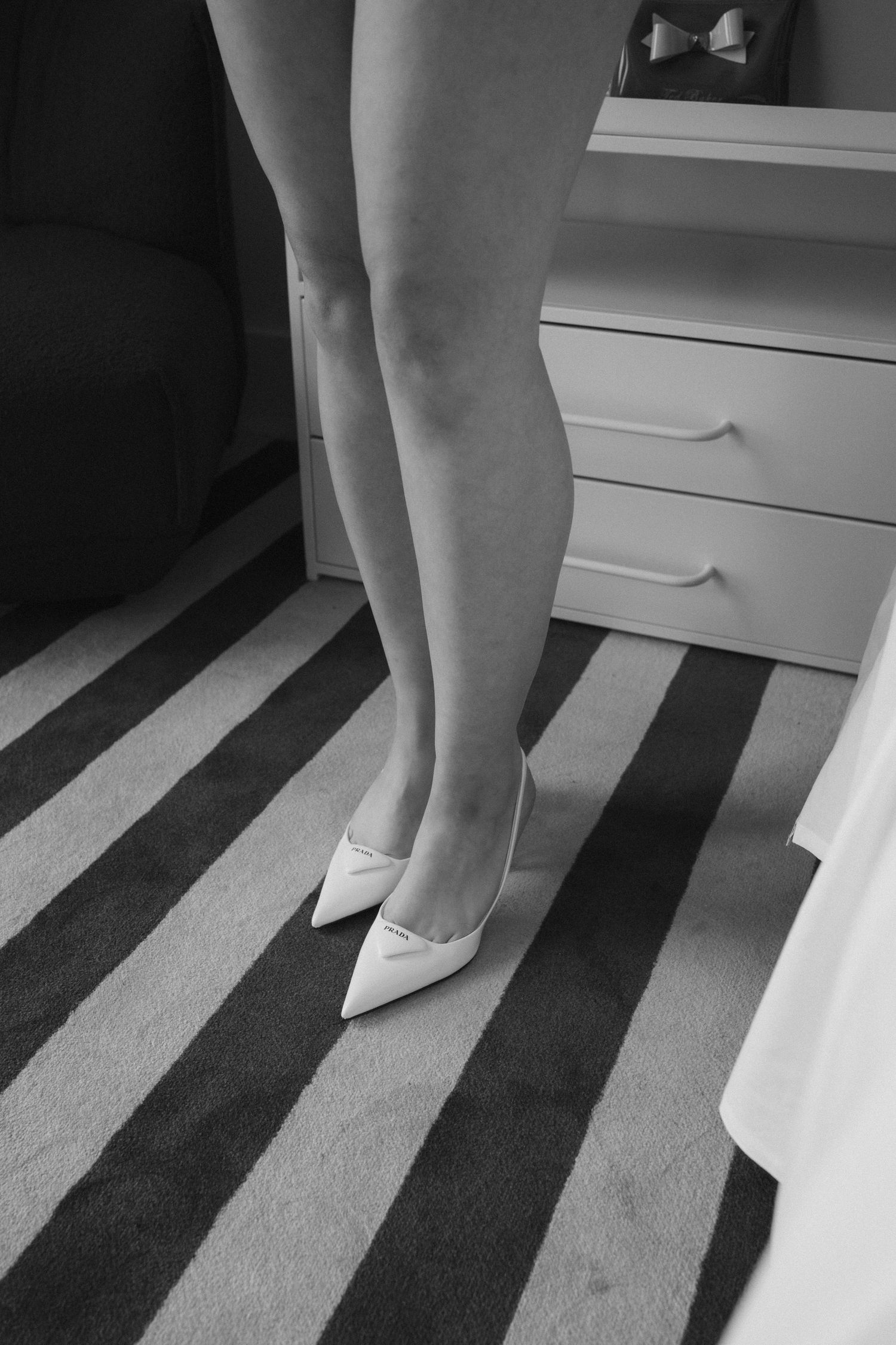 Black and white photo of womens legs wearing white pointed toe heels standing on striped carpet near dresser.