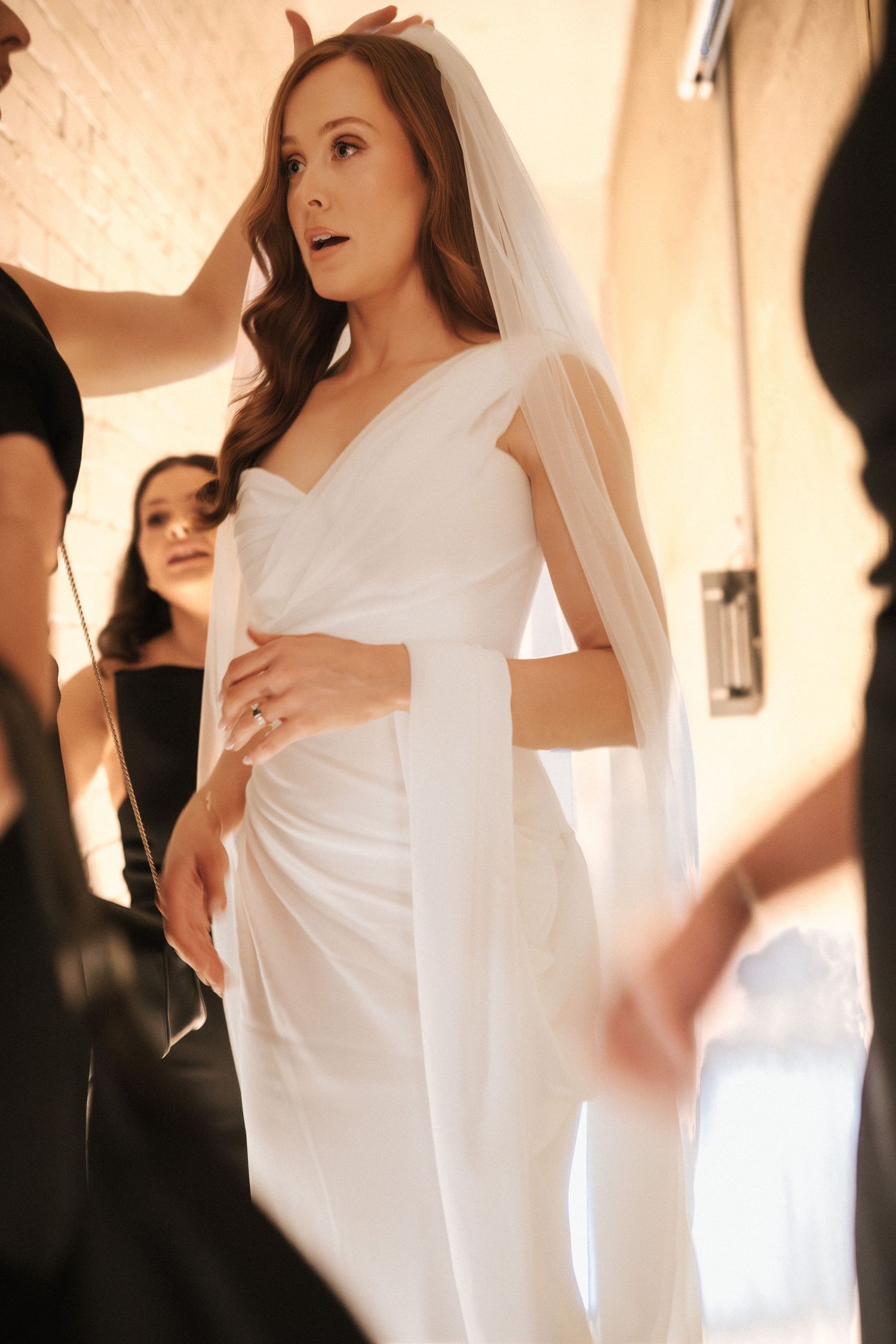 Bride in white wedding gown and veil being assisted by bridesmaids in black dresses during bridal preparations.