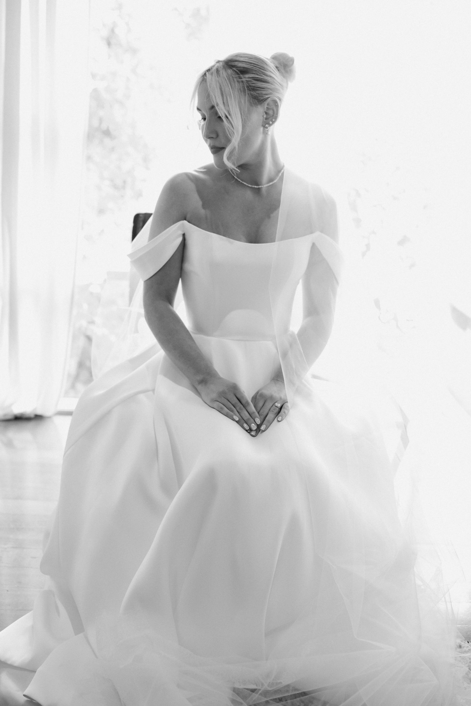 A bride in a classic white ballgown style wedding dress sits gracefully by a bright window in an editorial style photograph
