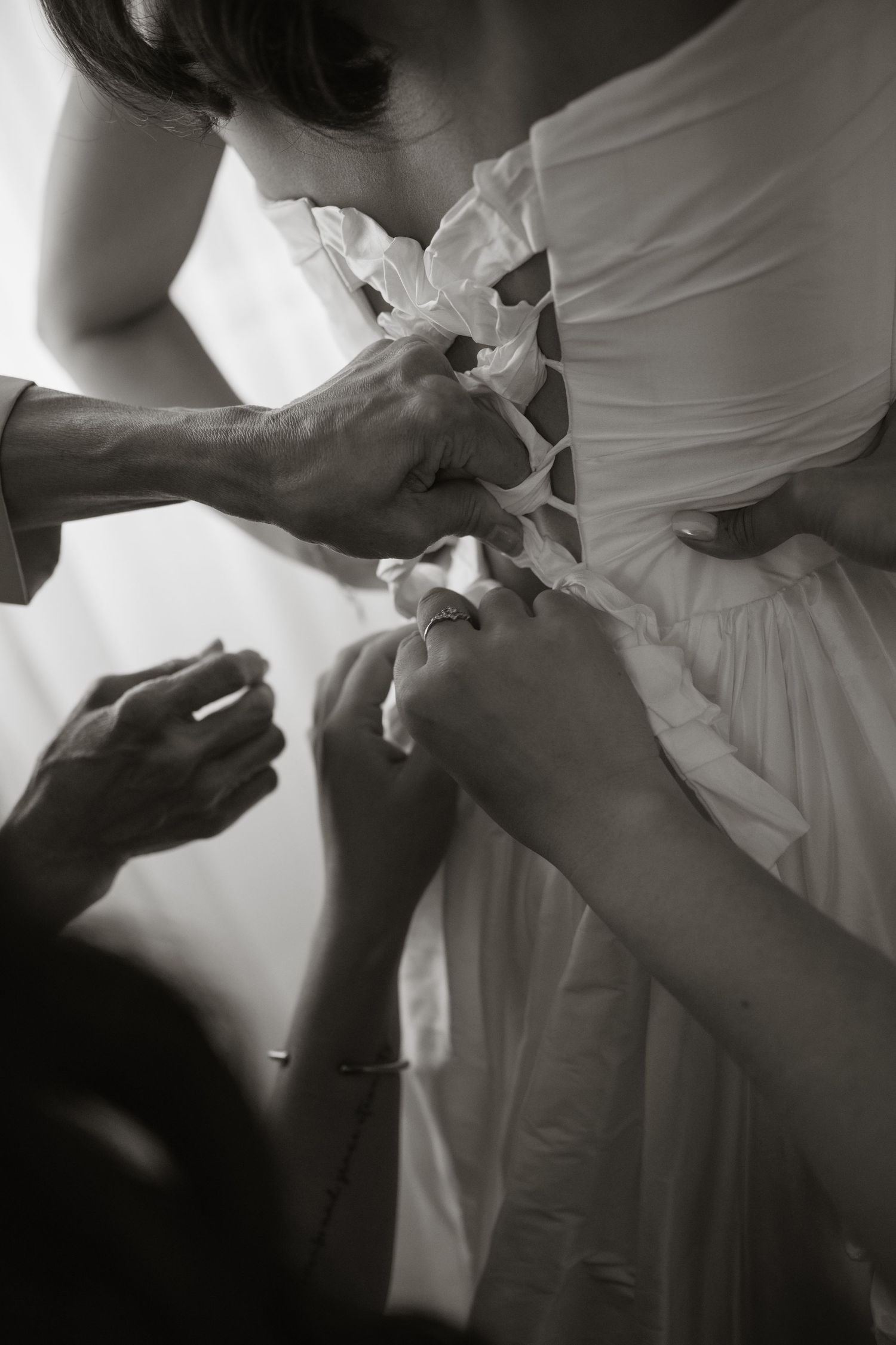 Helpers lacing up the back of a brides white wedding dress in black and white photo.