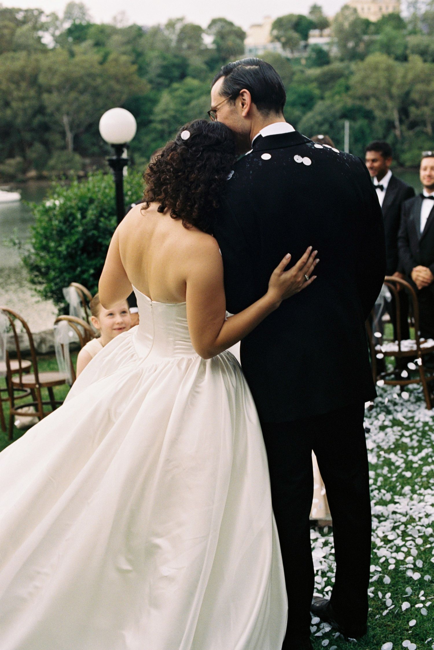 A bride in a white wedding dress and groom in black tuxedo embrace in a garden filled with white flowers.