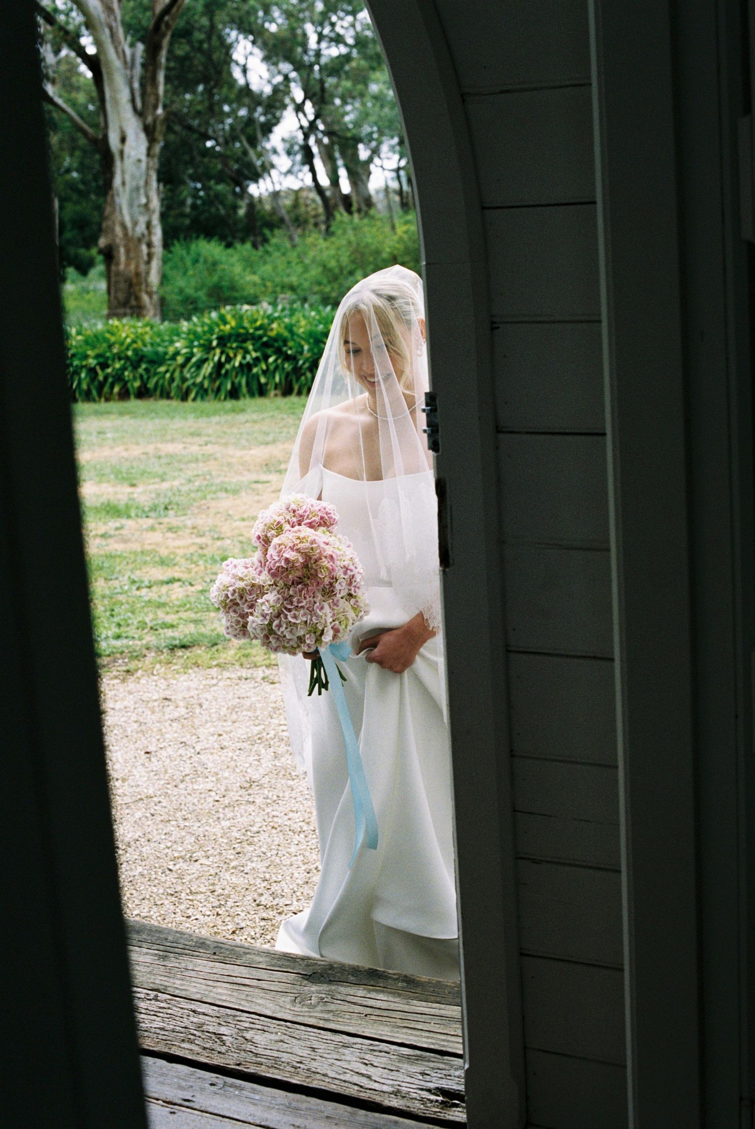 Wedding scene captured through doorway showing bride with bouquet and flowing dress.