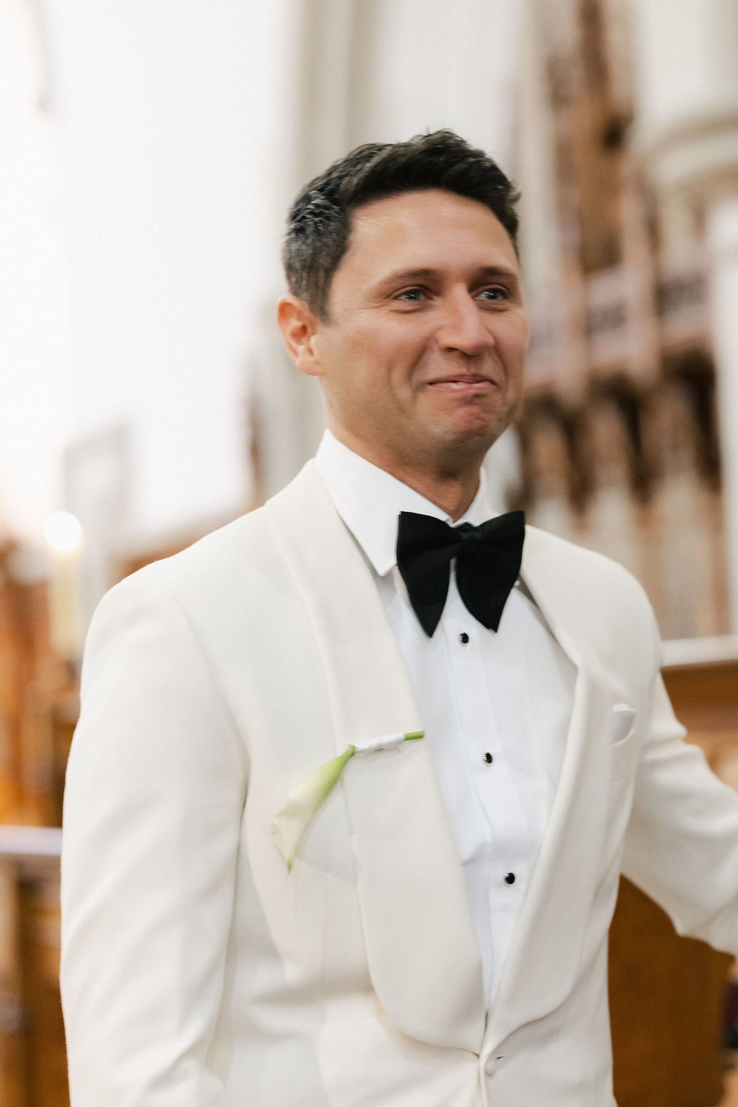 Groom in classic white tuxedo and black bow tie smiling at wedding.