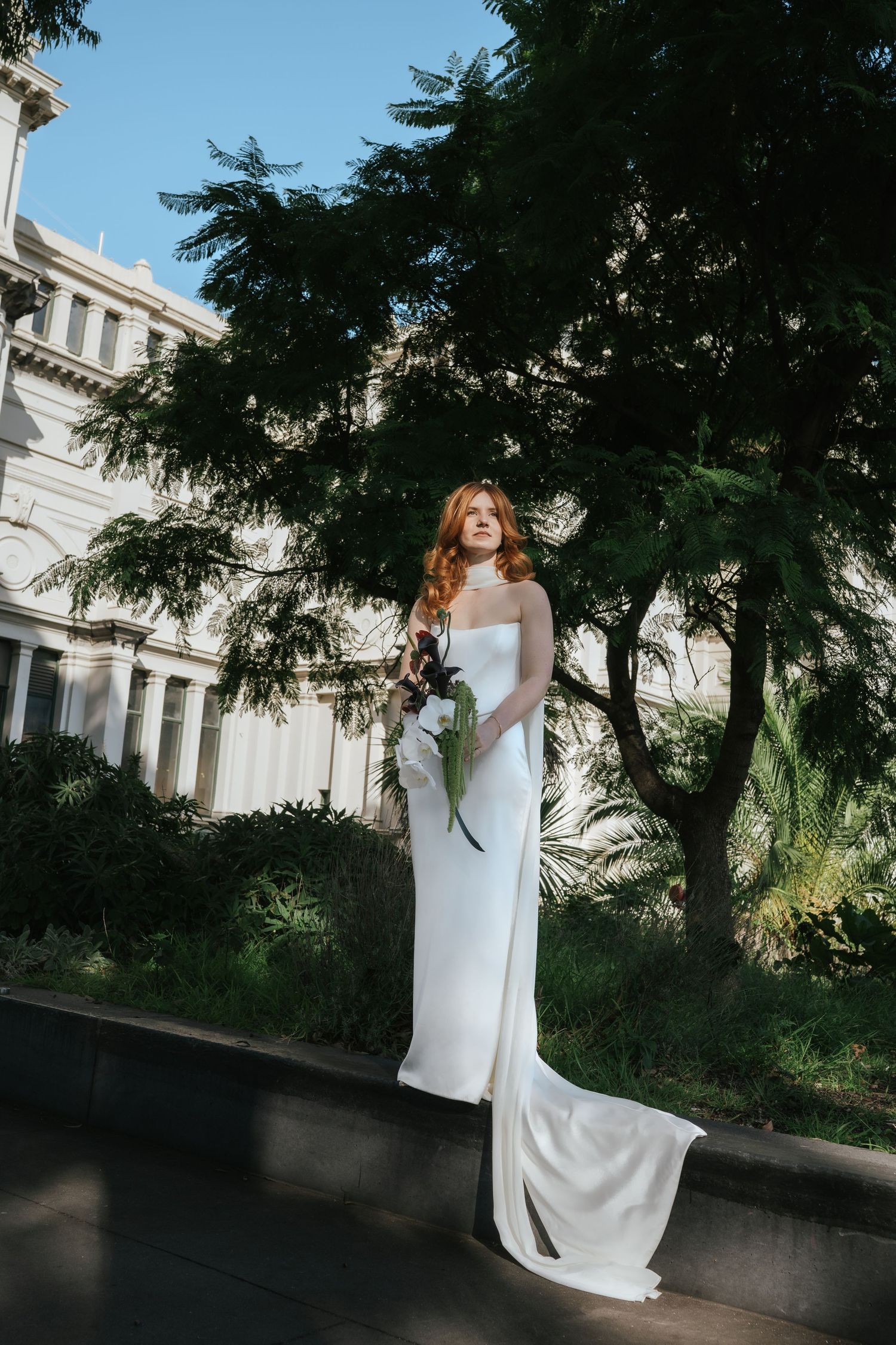 Elegant bride in white strapless gown holding bouquet, standing near lush green trees and classical building.