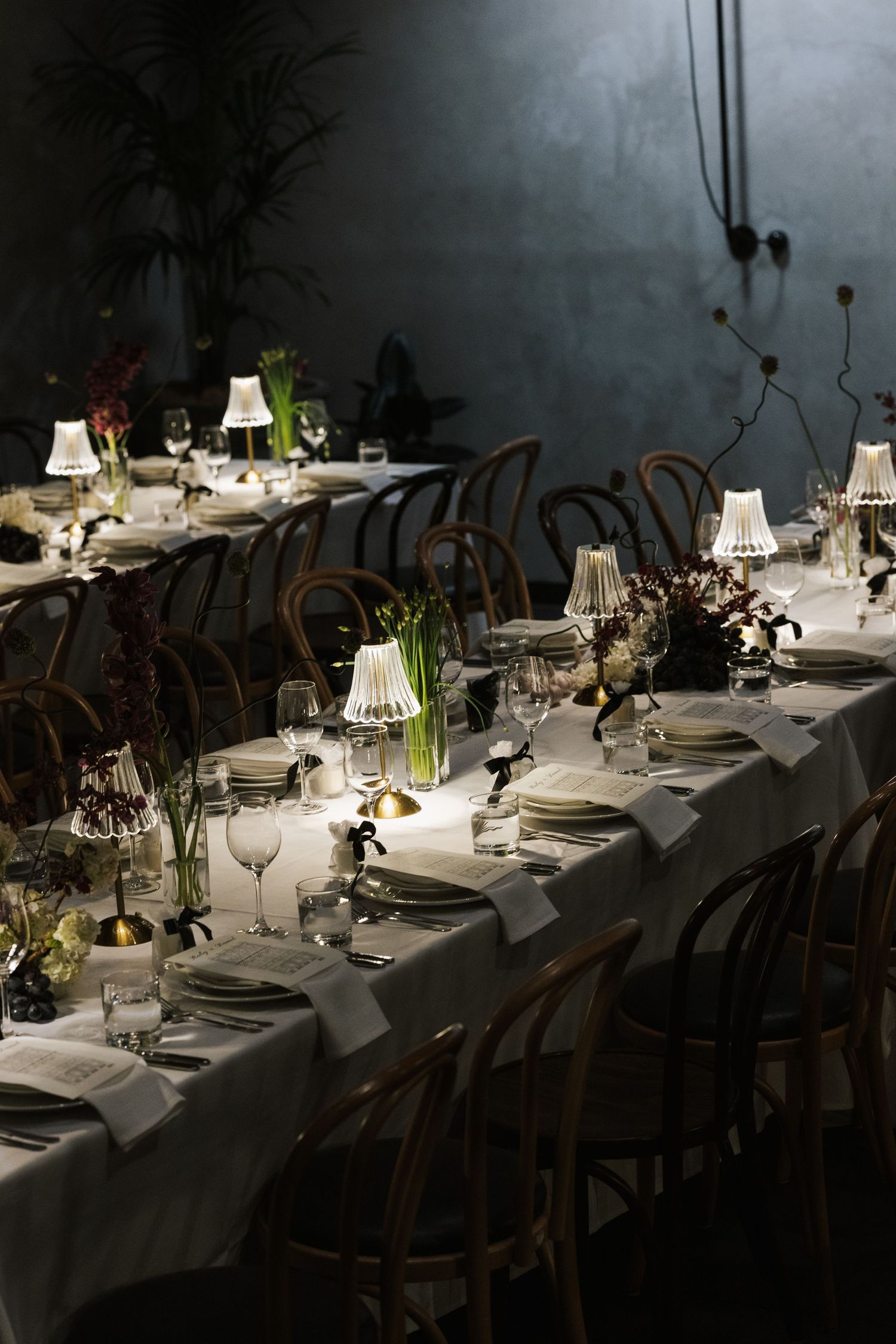 Elegant long dining table set with white linens, small lampshades, glassware, and floral arrangements in a moody venue.