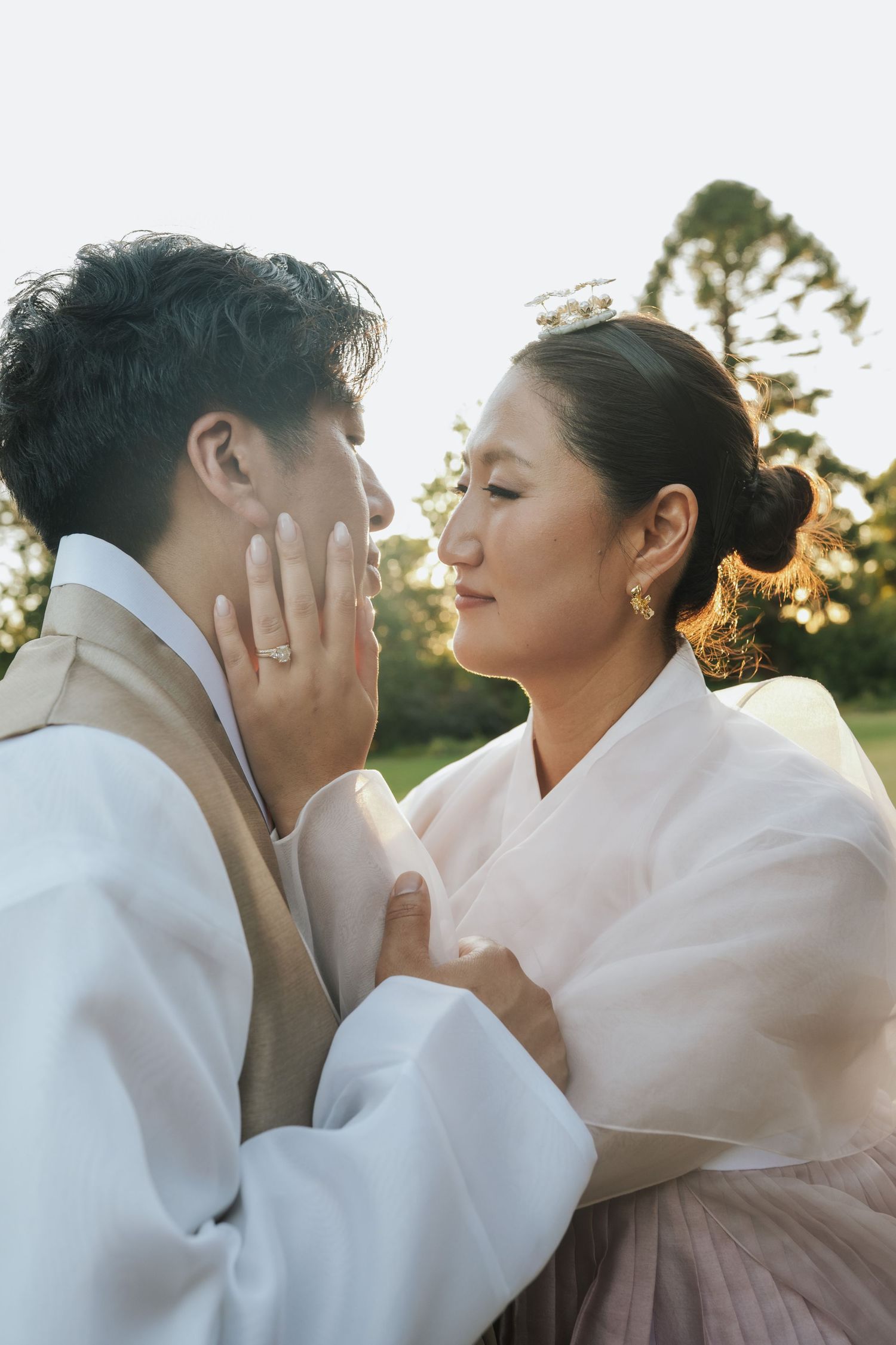 A couple in white traditional Korean hanbok outfits share an intimate moment at sunset in a garden setting.