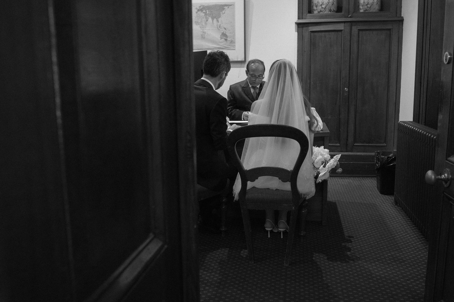 Black and white photo of a bride and groom signing wedding documents at a table with an officiant.