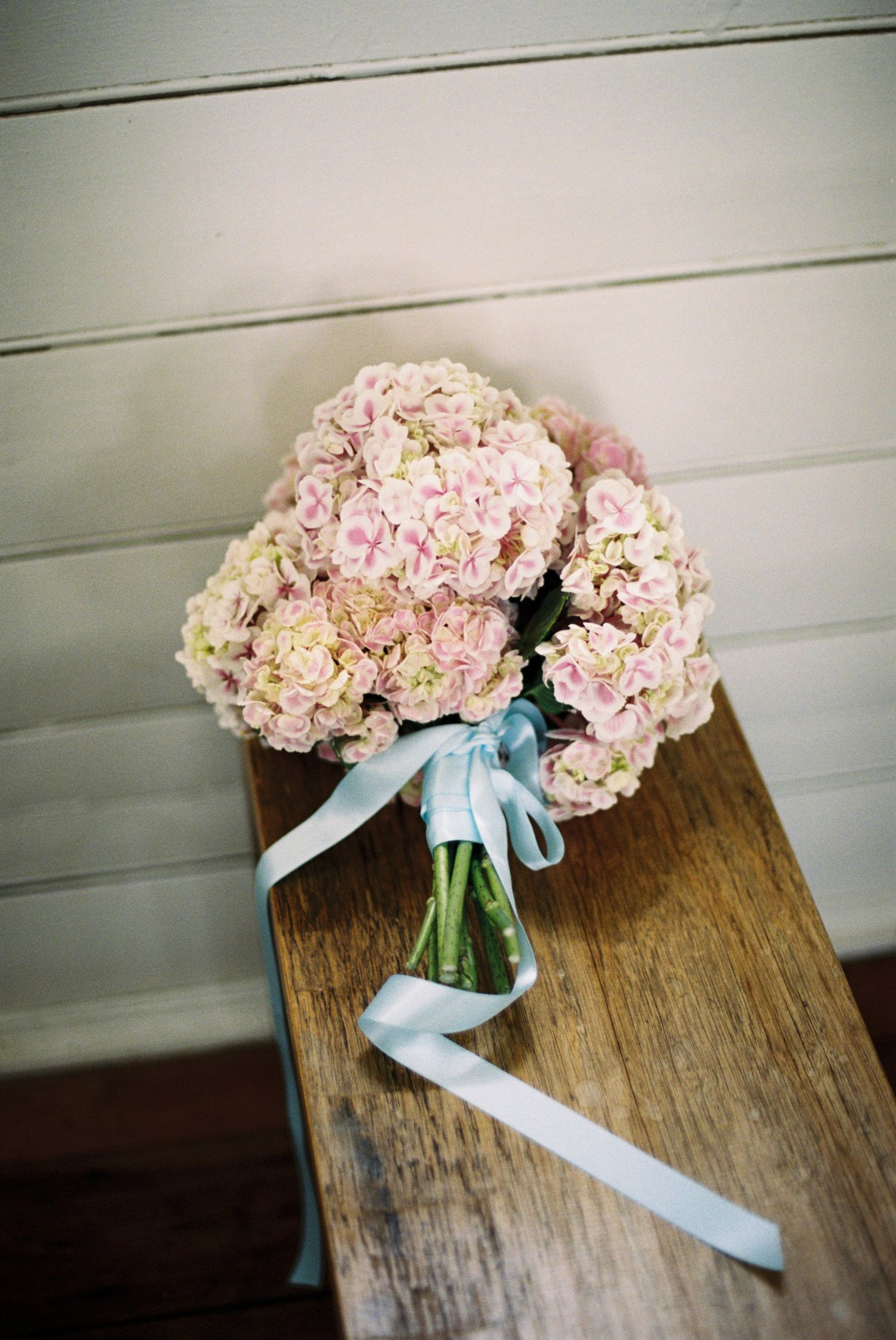 Pink hydrangea bouquet with blue ribbon sitting on rustic wooden surface.