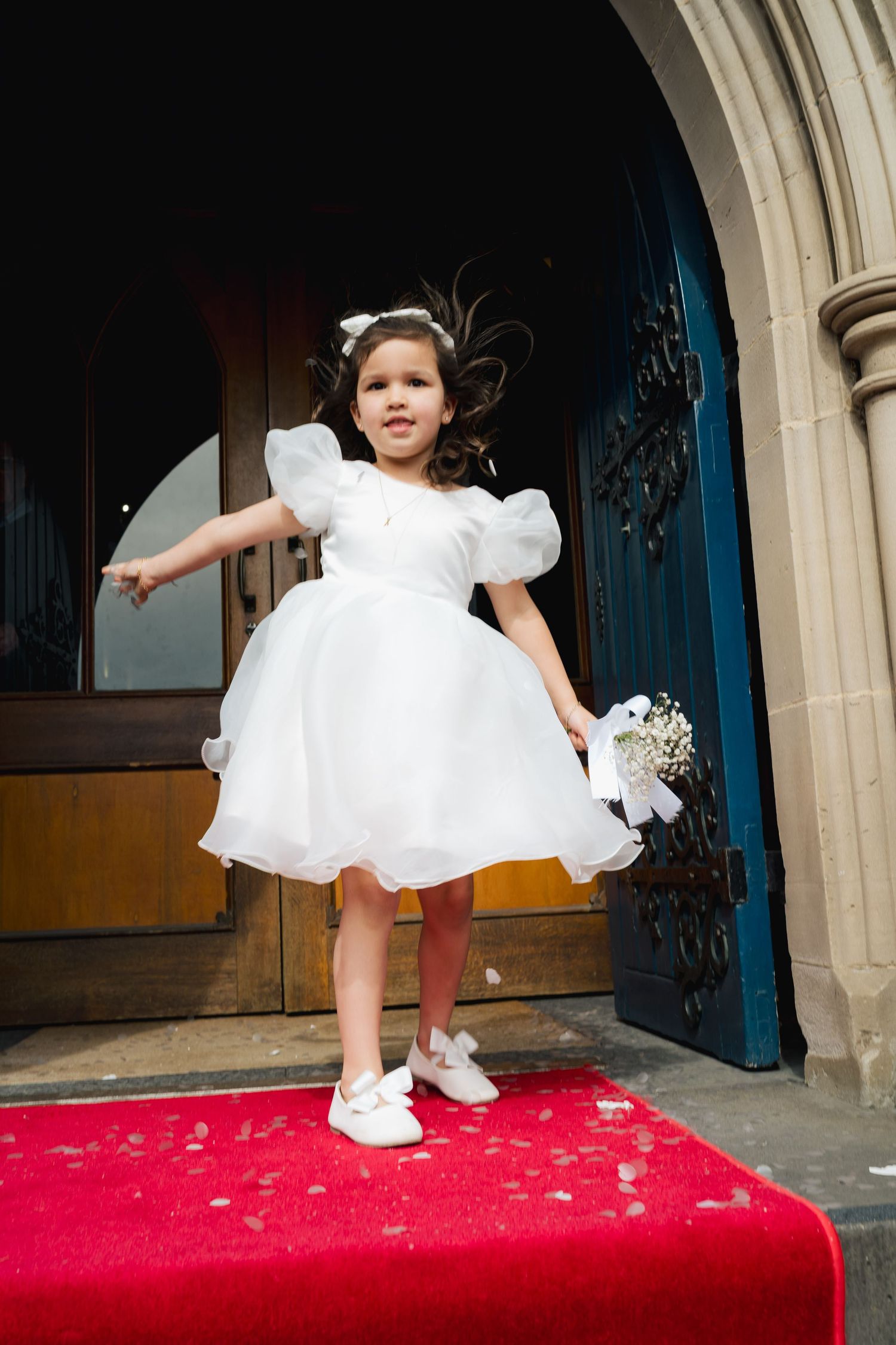 A young flower girl in a white dress jumps playfully on a red carpet in front of an ornate doorway.