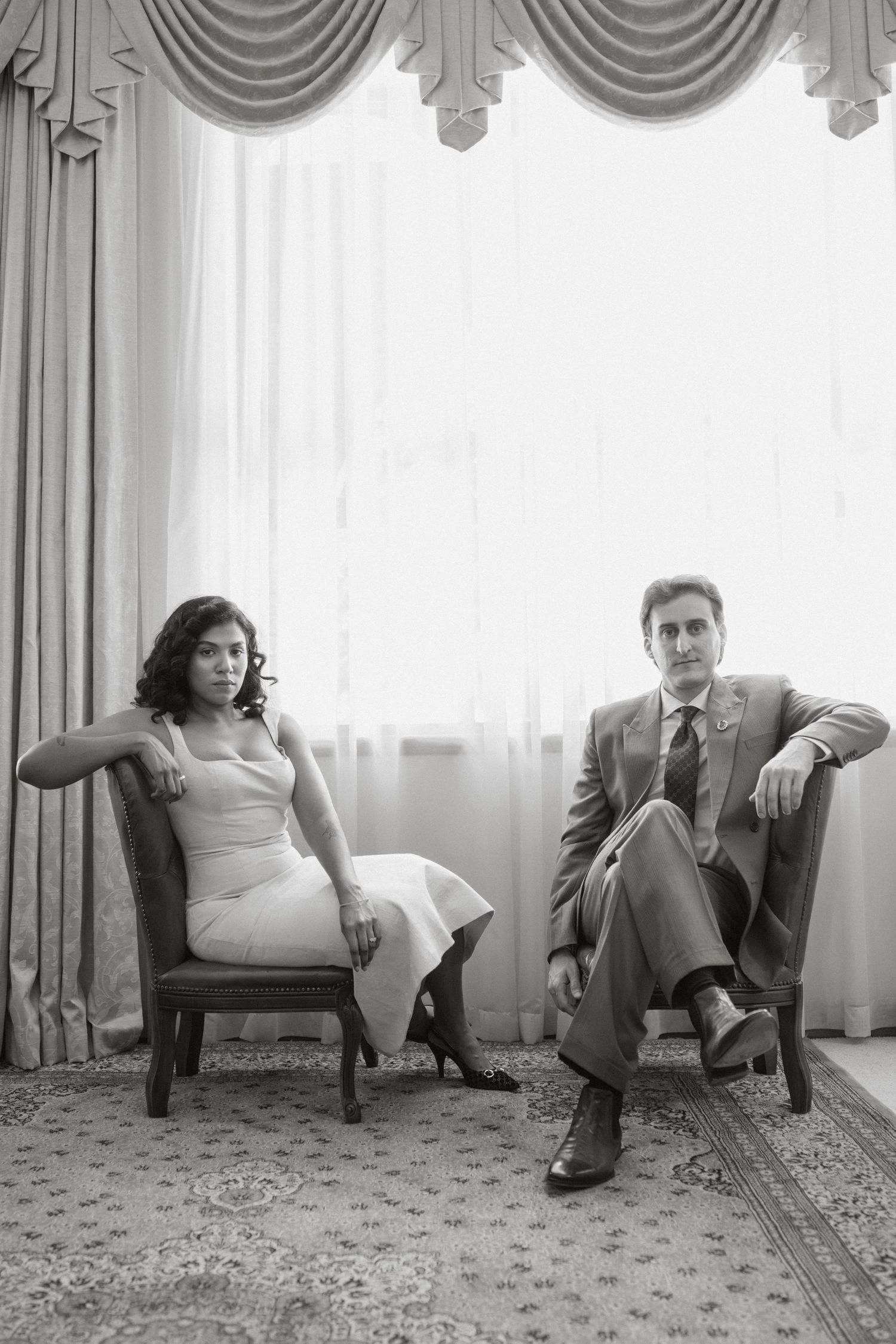 Black and white portrait of a man in suit and woman in dress seated in armchairs by elegant curtained window.