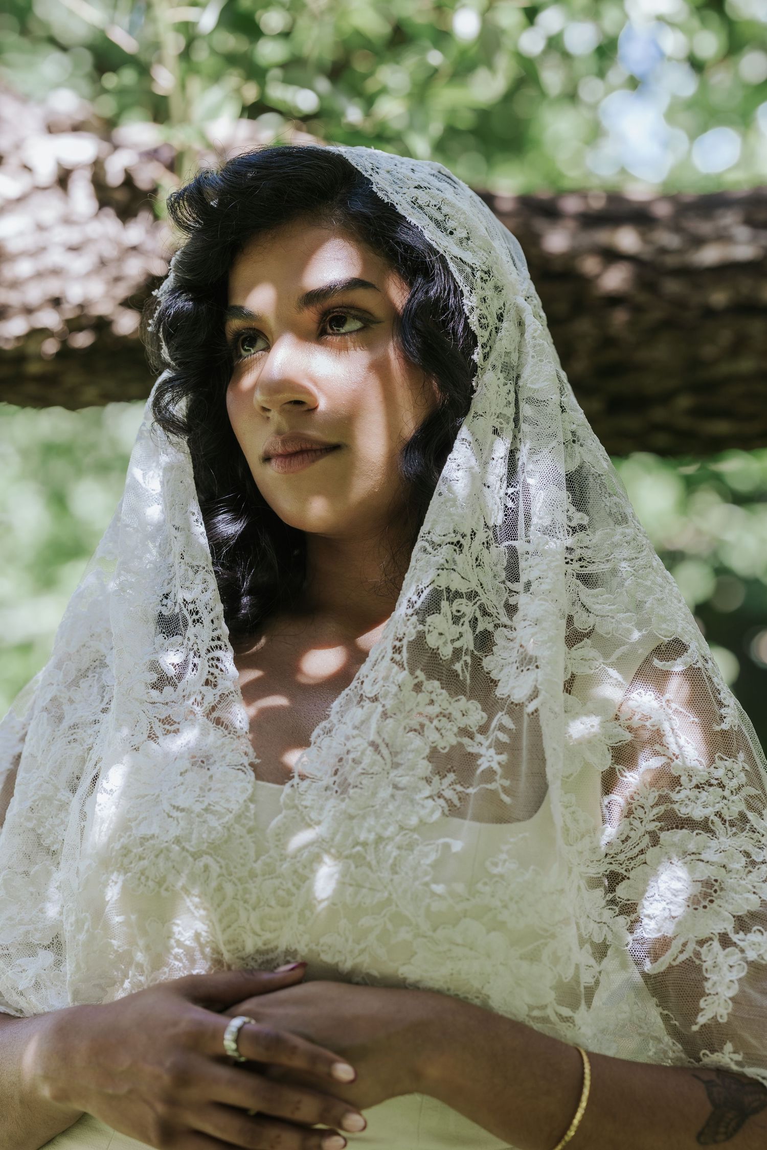 A bride in a lace veil and wedding dress poses gracefully outdoors under a tree in dappled sunlight.
