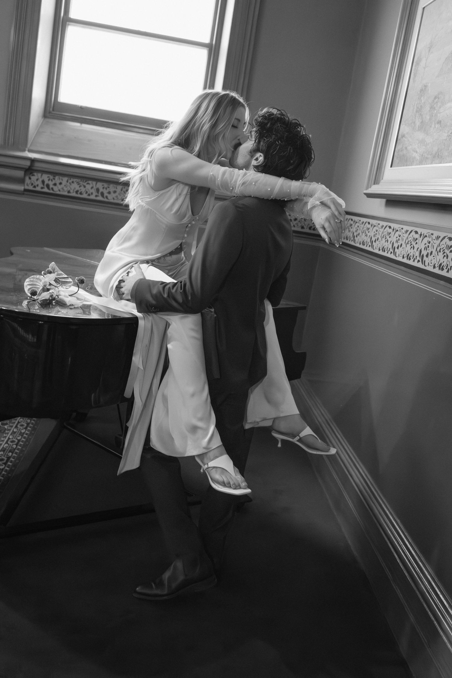 Black and white photo of a couple sharing a romantic kiss, with the man lifting the woman in a warmly lit room.
