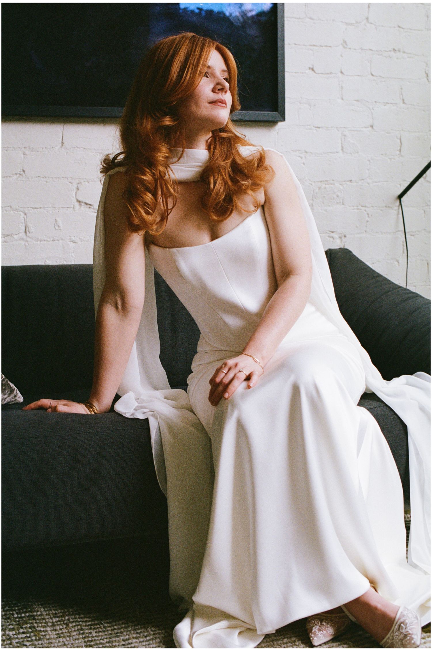 Elegant bride in white satin gown with sheer cape sits gracefully on dark sofa.