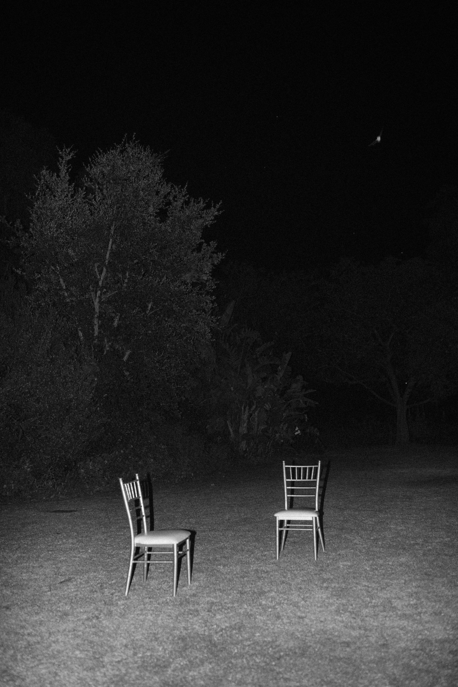 Two empty chairs on a dark lawn at night with trees in the background, black and white photography.