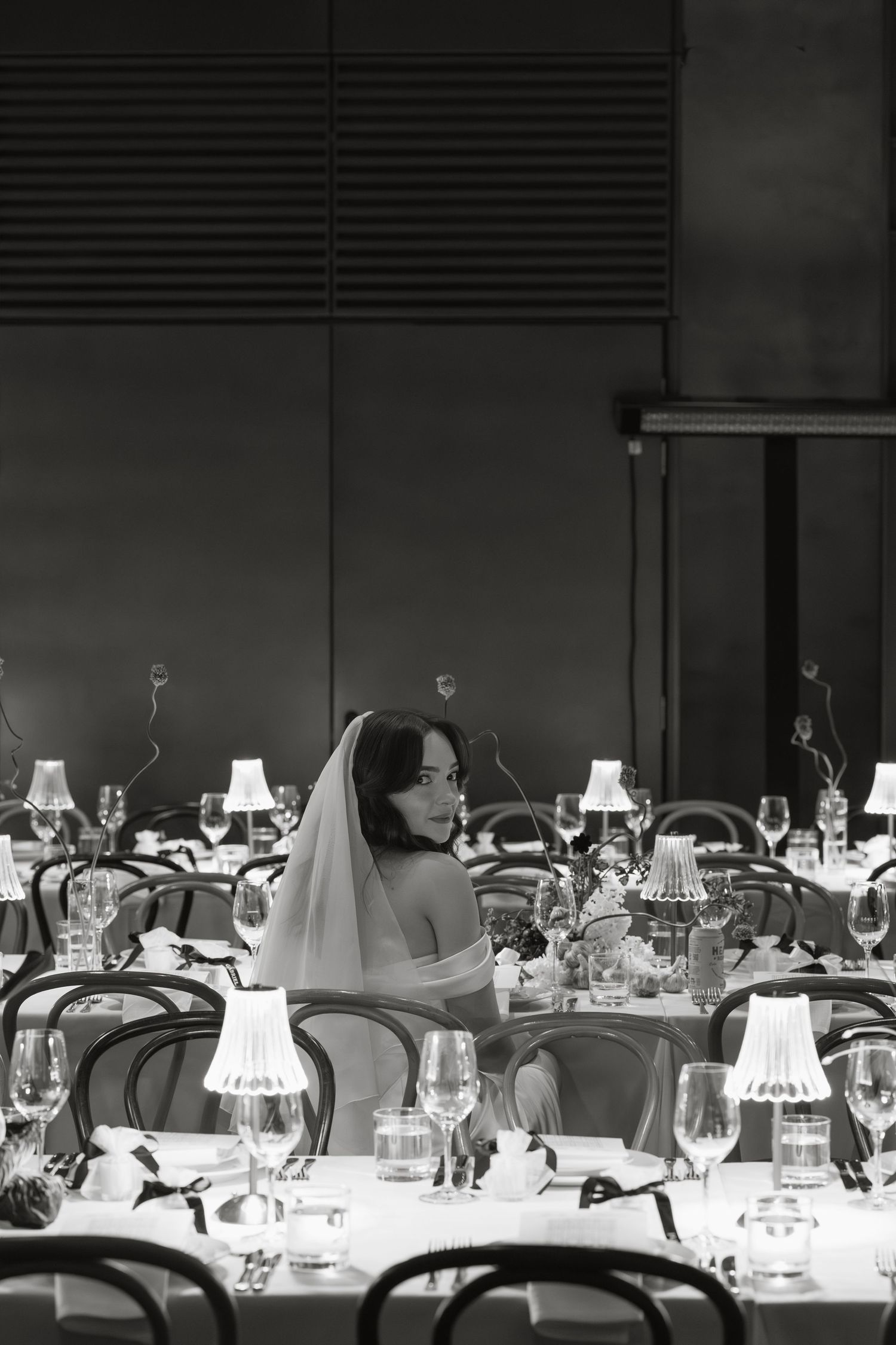 Bride in a veil sits alone at an elegantly set banquet table in a dimly lit reception hall.