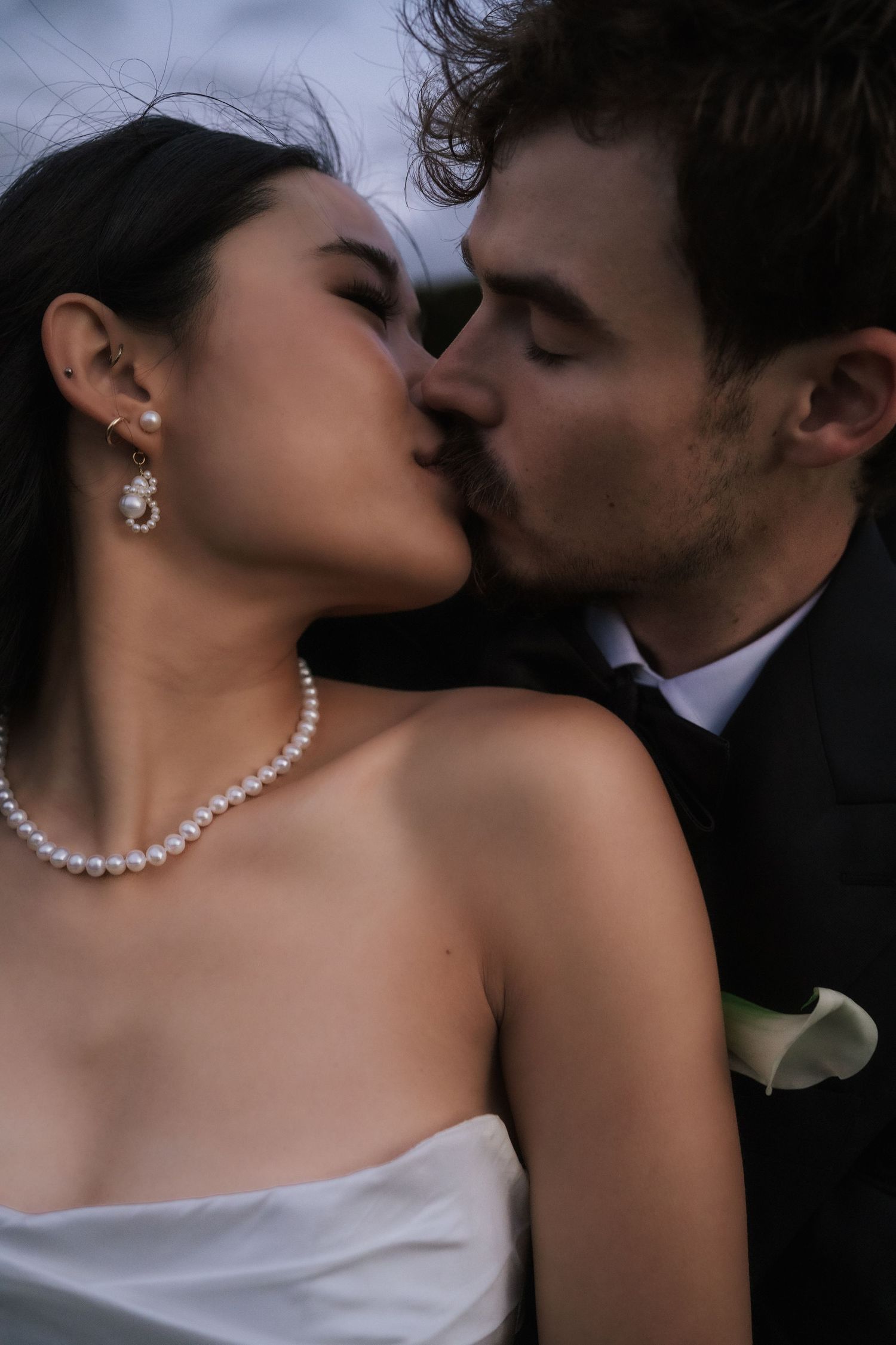 A bride wearing a pearl necklace and groom in a black suit share a romantic kiss at their wedding.