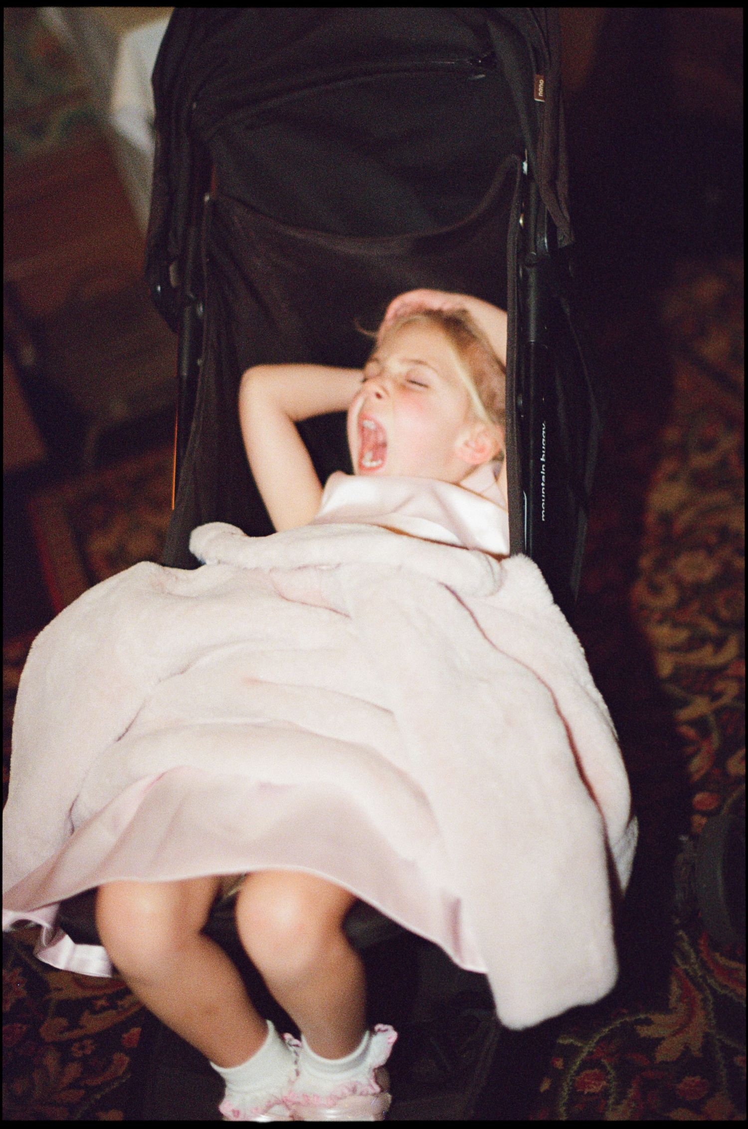 Tired toddler girl in pink dress yawning with arms raised while sitting in black stroller.