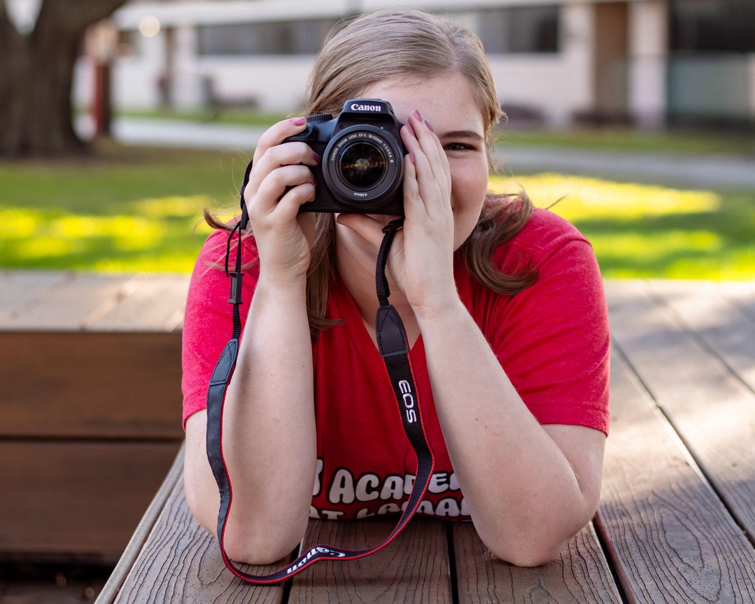 Young woman in red t-shirt photographing with a Canon DSLR camera outdoors on a wooden bench.