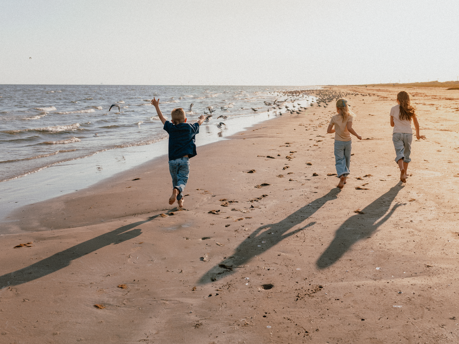 Three children running joyfully along a sandy beach shore at sunset, casting long shadows on the sand.
