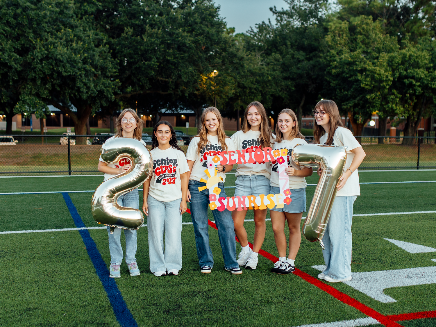 Six girls pose on a football field holding gold balloon numbers 2 and 7, wearing matching shirts for a senior celebration.