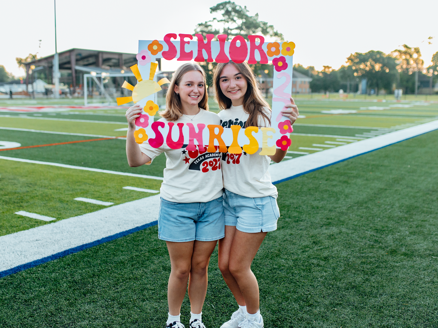 Two smiling senior girls hold a colorful decorative frame reading Senior Sunrays on a football field.