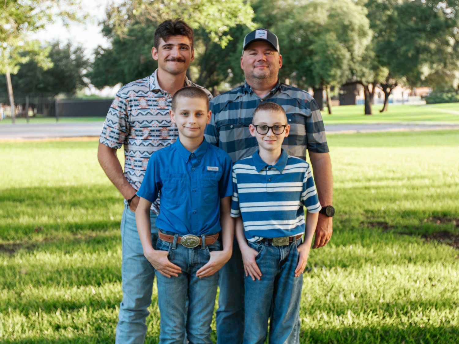 Family portrait of four males, two adults and two boys, smiling outdoors on green grass in a park setting.