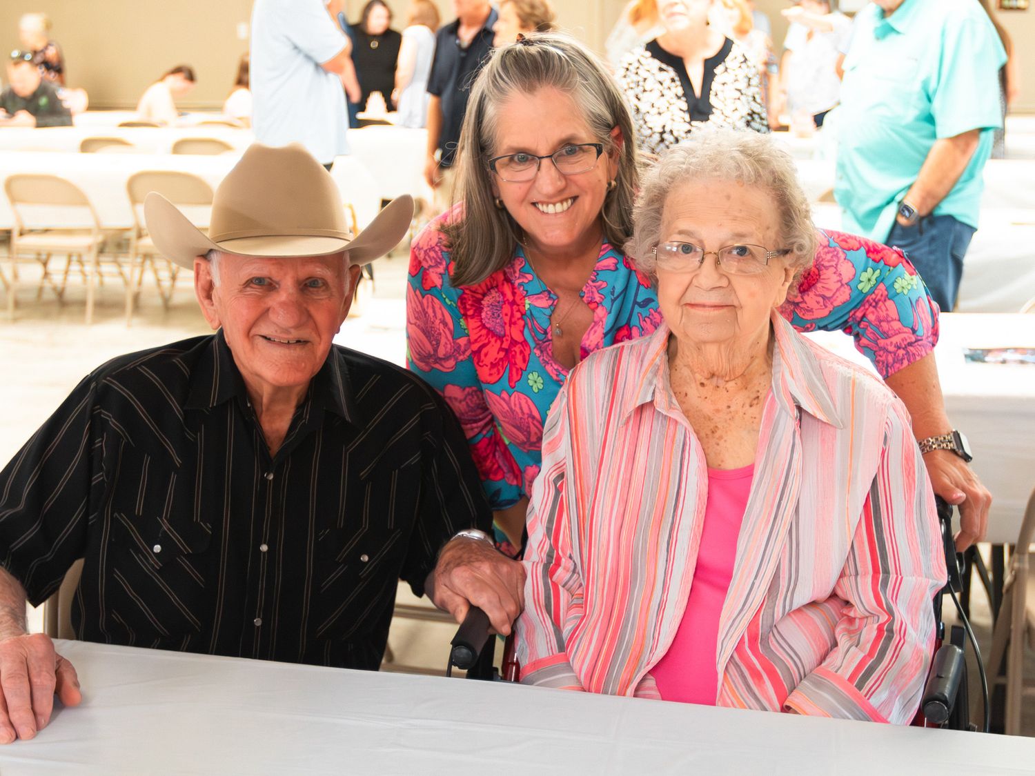 Elderly couple sitting at a table with a smiling woman behind them at an indoor gathering event.