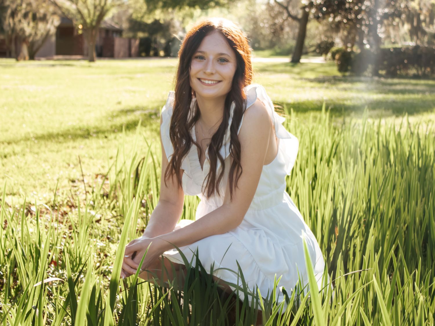Young woman in white dress smiling while crouching in lush green grass outdoors on a sunny day.