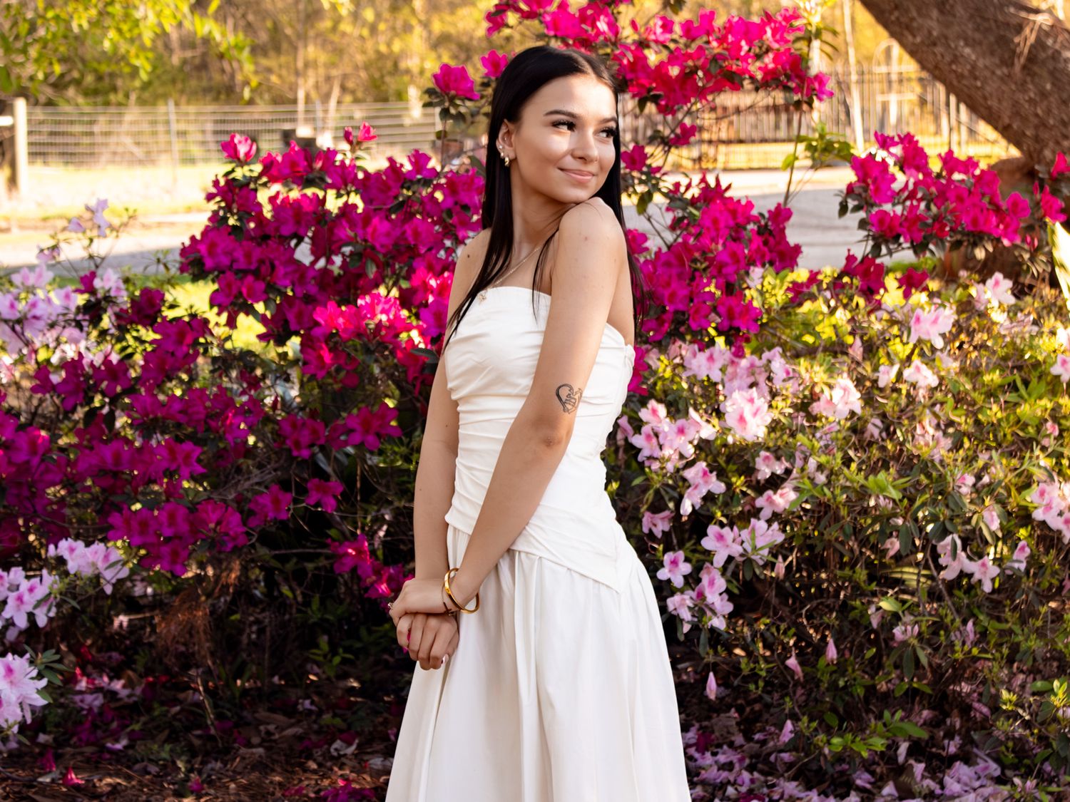 Woman in white dress posing before vibrant pink and purple azalea bushes in a sunny garden.