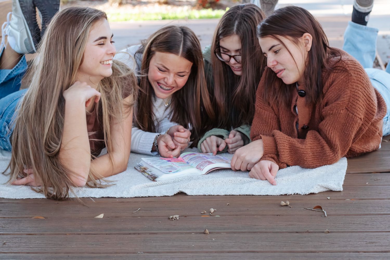 Four teenage girls lying on a wooden deck, smiling and reading a book together outdoors.
