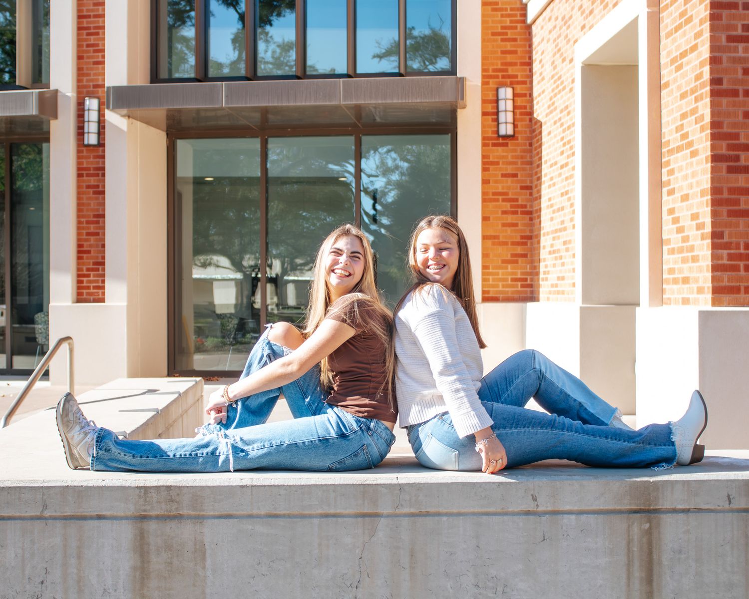 Two smiling young women sitting back-to-back on a concrete ledge outside a brick building, wearing jeans.