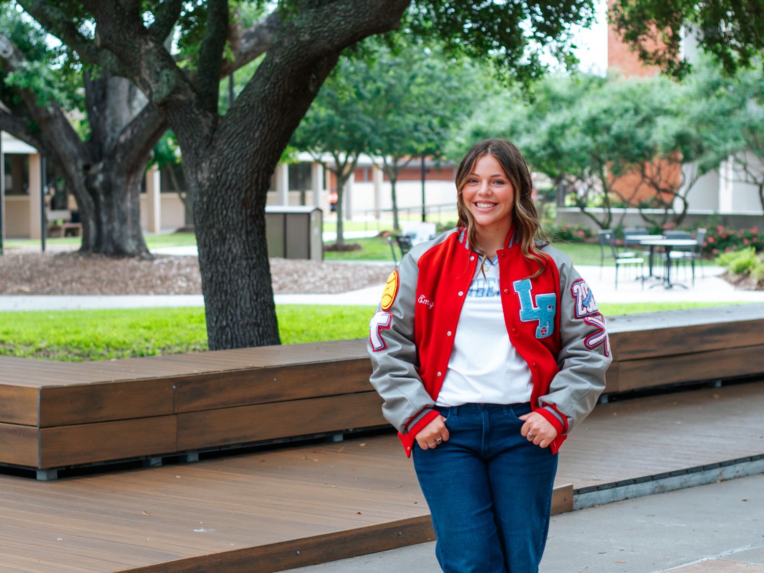 Young woman in red varsity jacket smiling on a college campus with trees and benches in background.