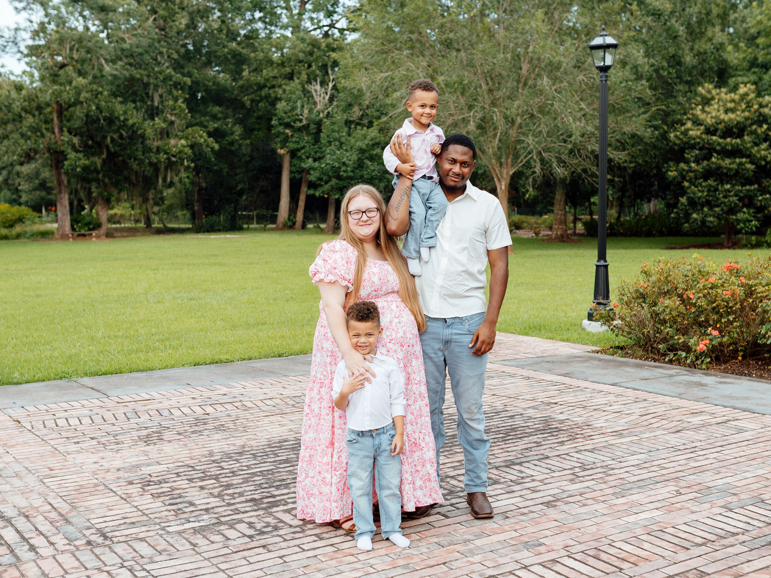 Happy interracial family of four posing outdoors on brick path surrounded by lush green trees and garden.