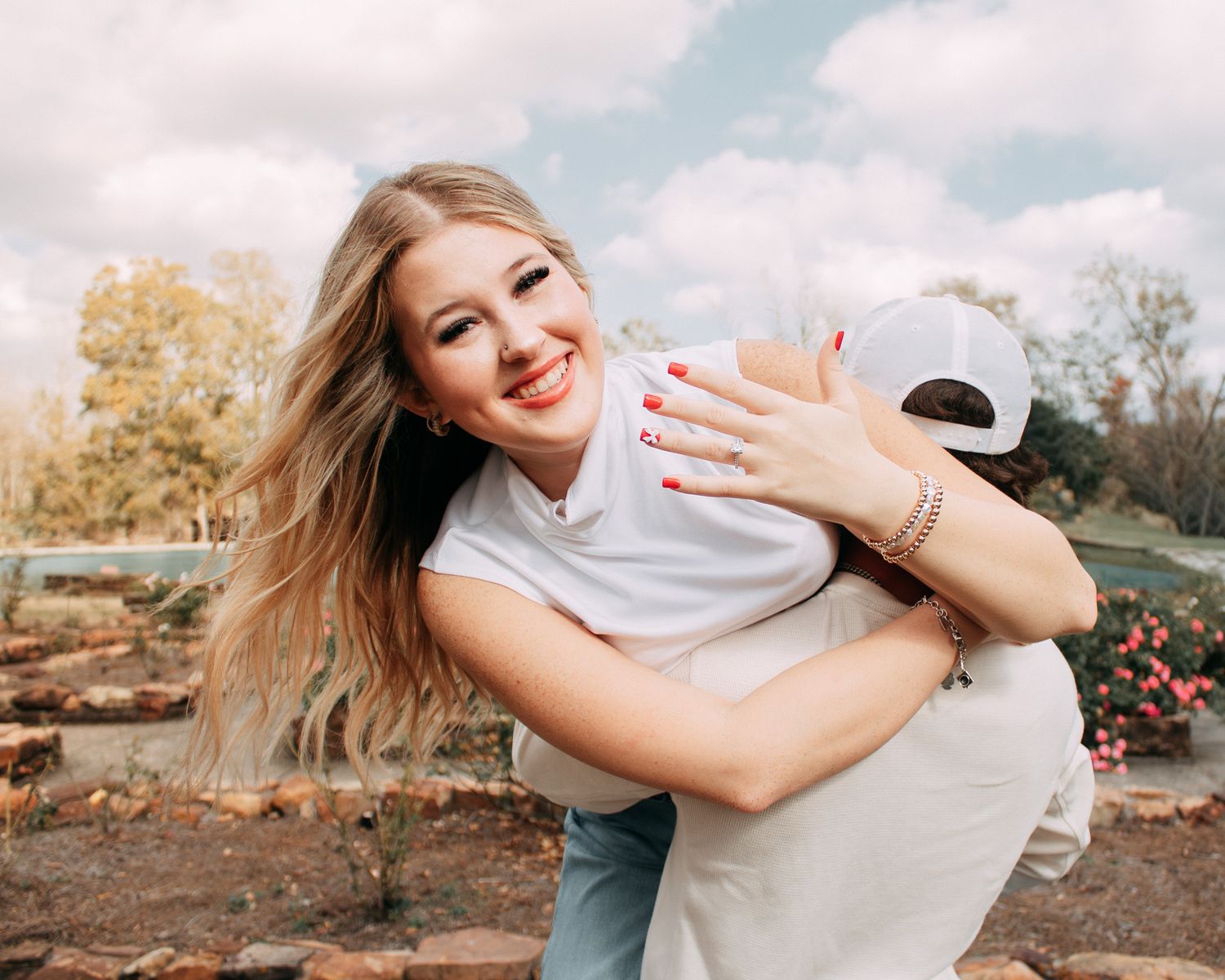 Smiling blonde woman with red nails hugging someone outdoors in a garden setting on a cloudy day.