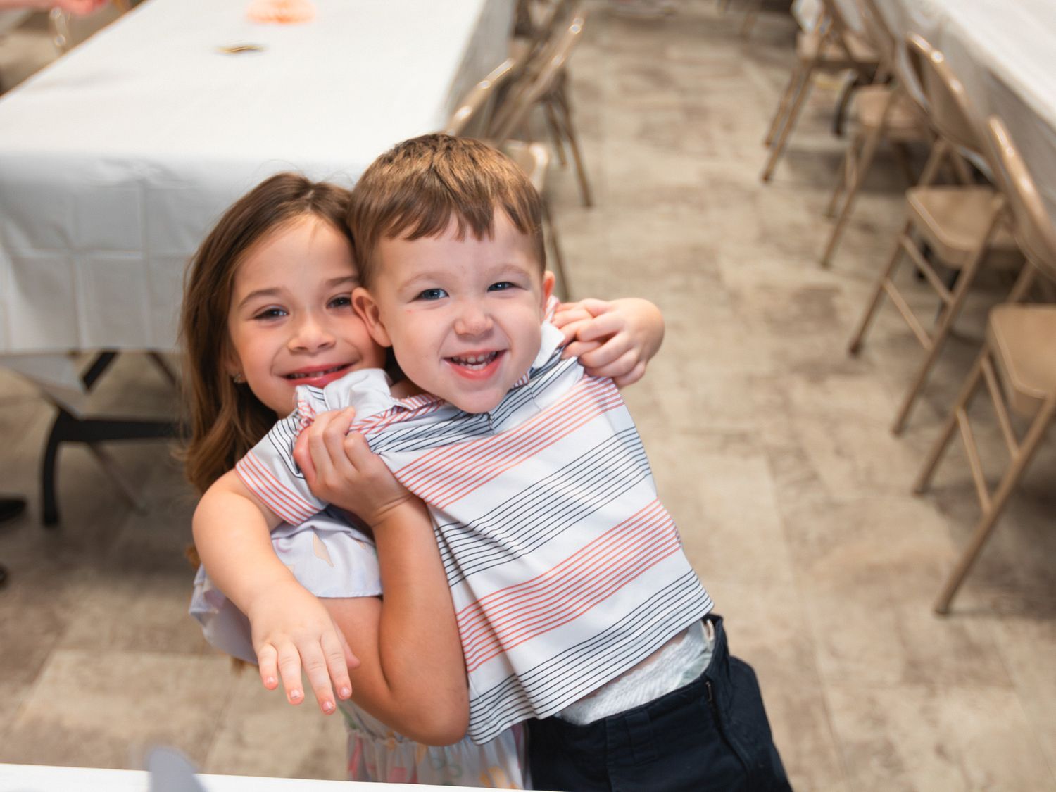Two young children hugging and smiling at a banquet hall with folding tables and chairs.