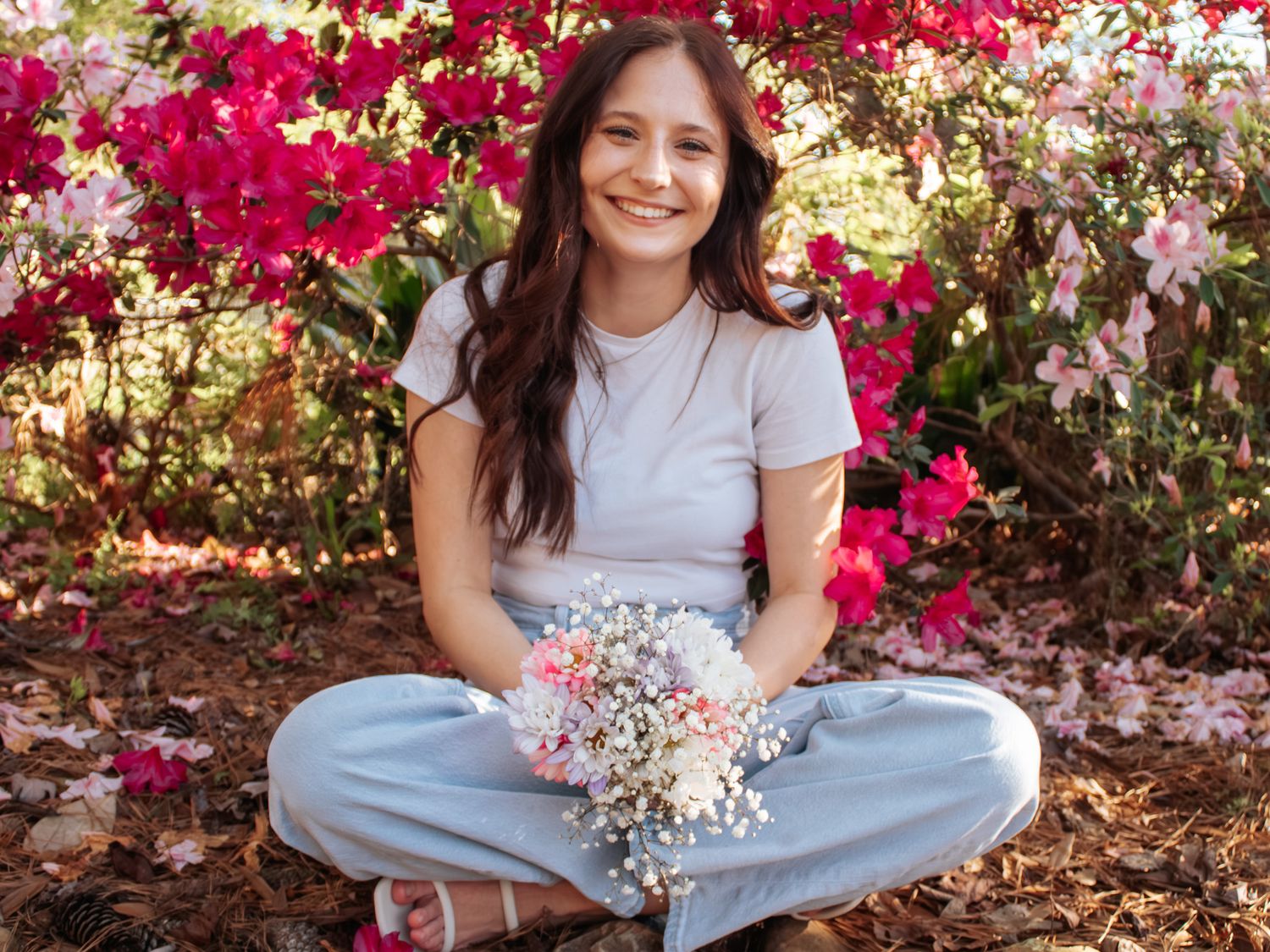 Young woman sitting cross-legged outdoors holding a bouquet of pink and white flowers with vibrant bougainvillea behind her.