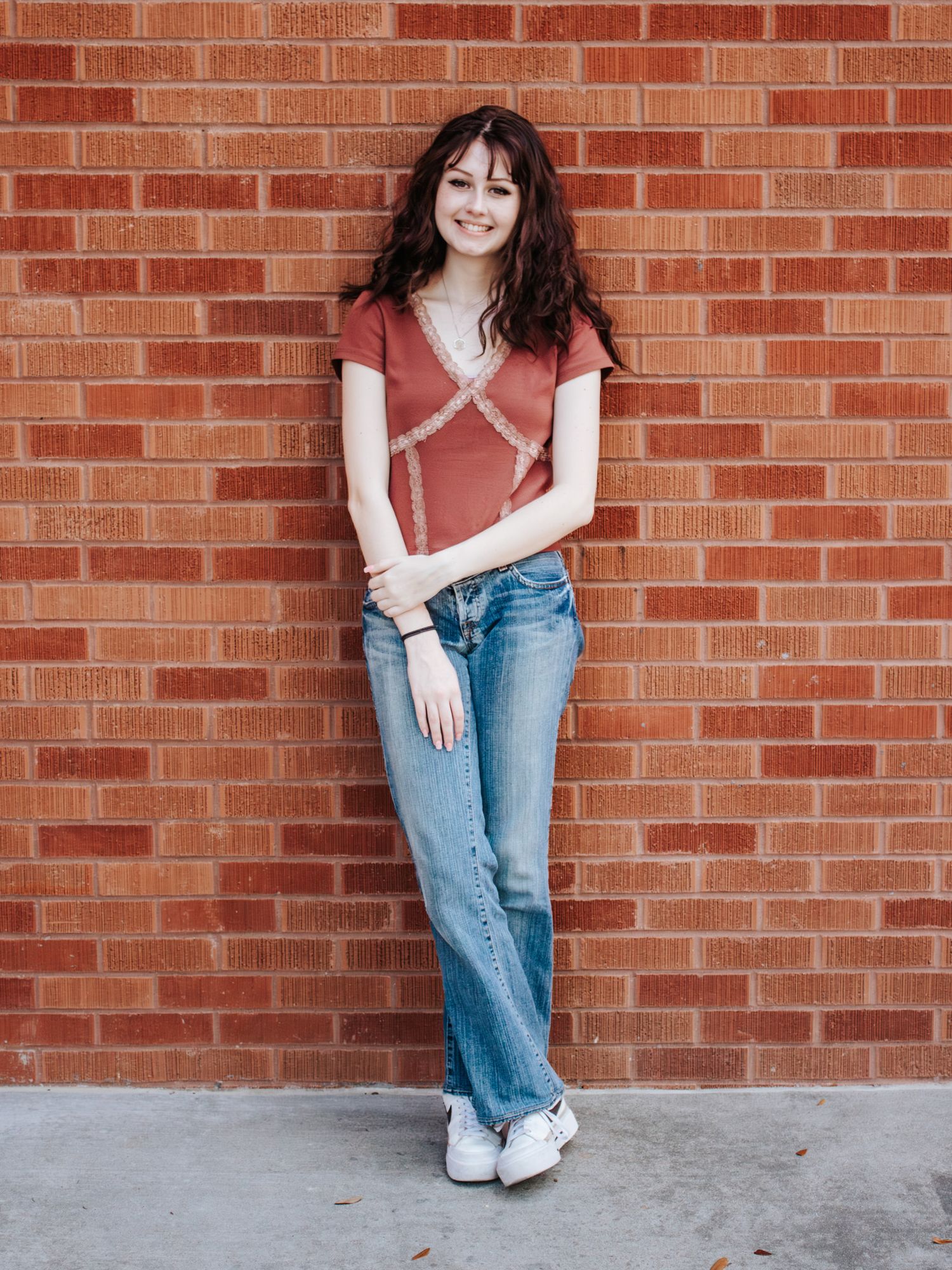 Young woman in rust top and flare jeans leaning against a brick wall, smiling casually.