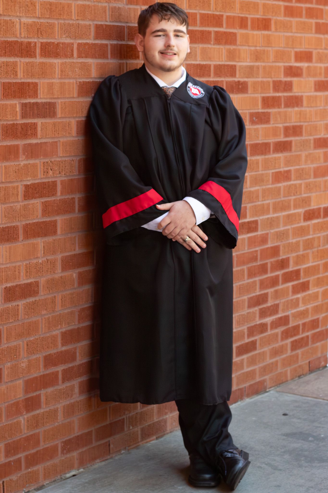 Young man in black and red graduation gown smiling against a brick wall.