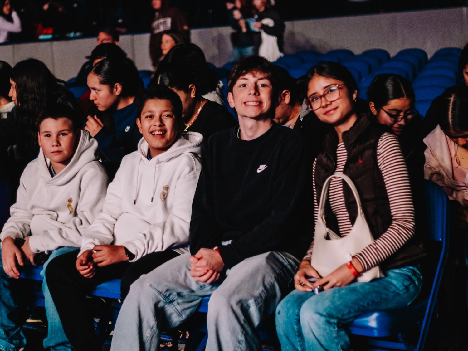 Four smiling teenagers sitting together in arena seats at a live event or concert.