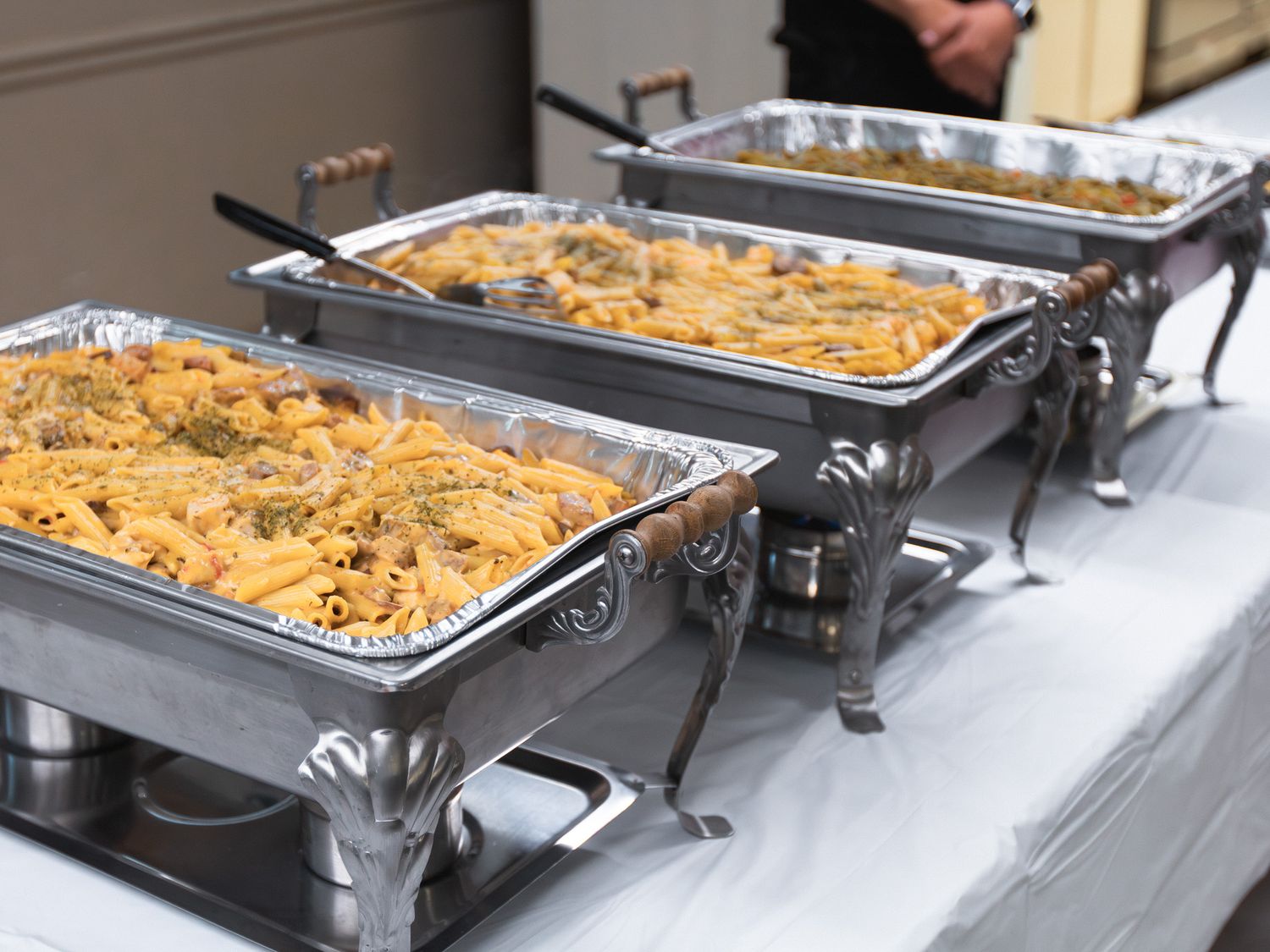 Three chafing dishes filled with pasta at a catered buffet event on a white tablecloth.