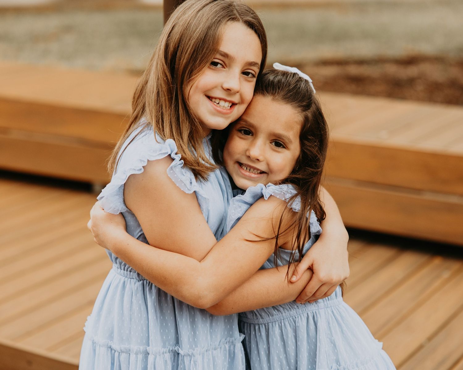 Two young girls in matching blue dresses hugging and smiling on a wooden deck.
