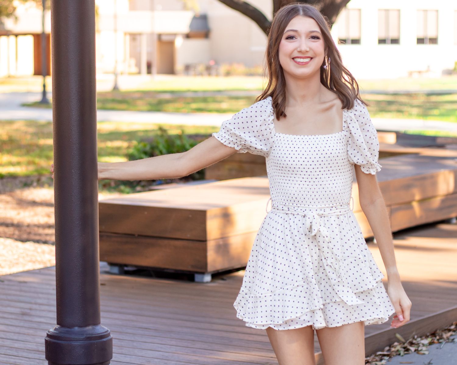 A person in a white polka dot dress poses outdoors near wooden benches and pillars in natural lighting.