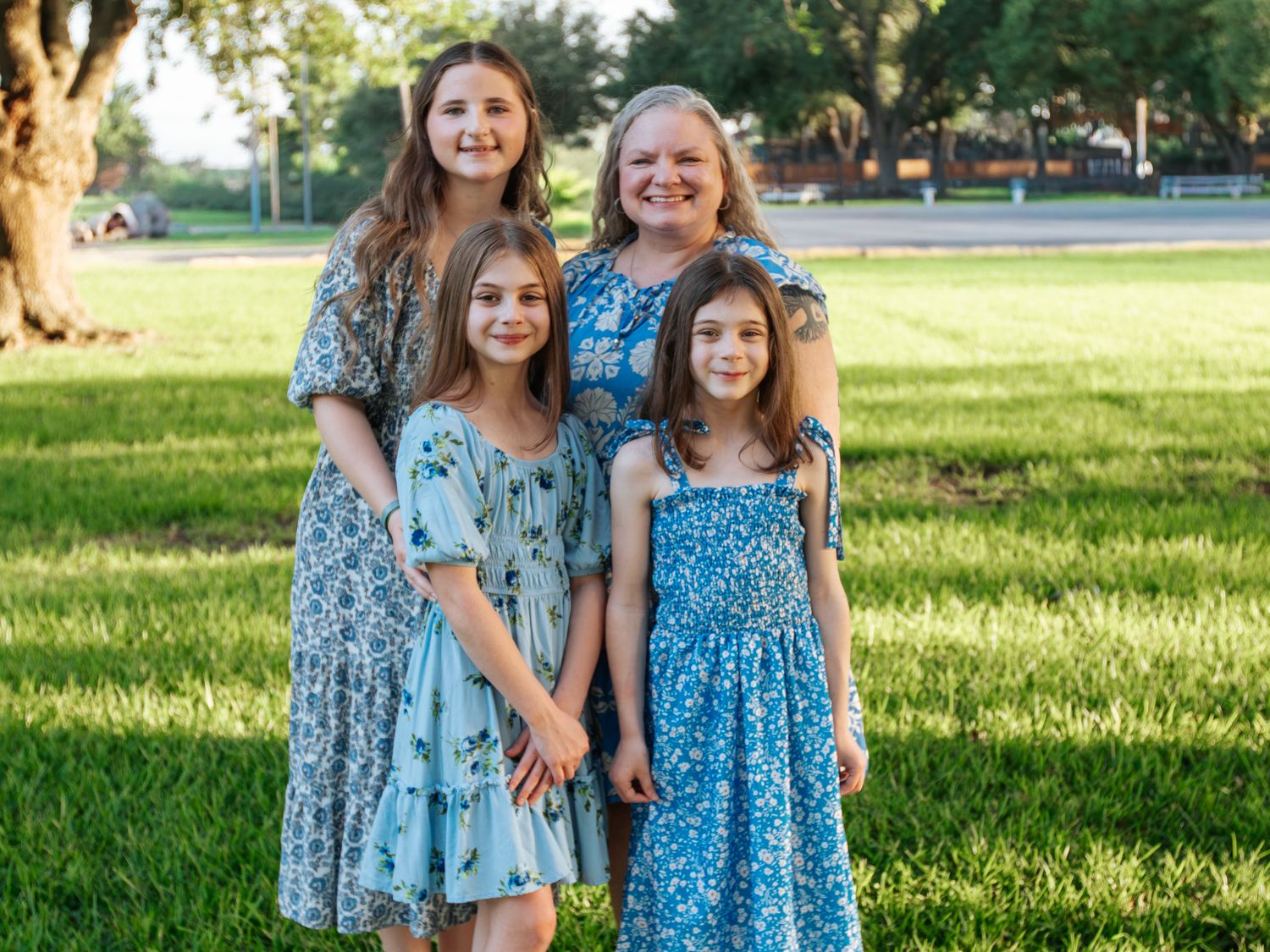 A woman and three girls in matching blue floral dresses smile together outdoors on a green lawn.