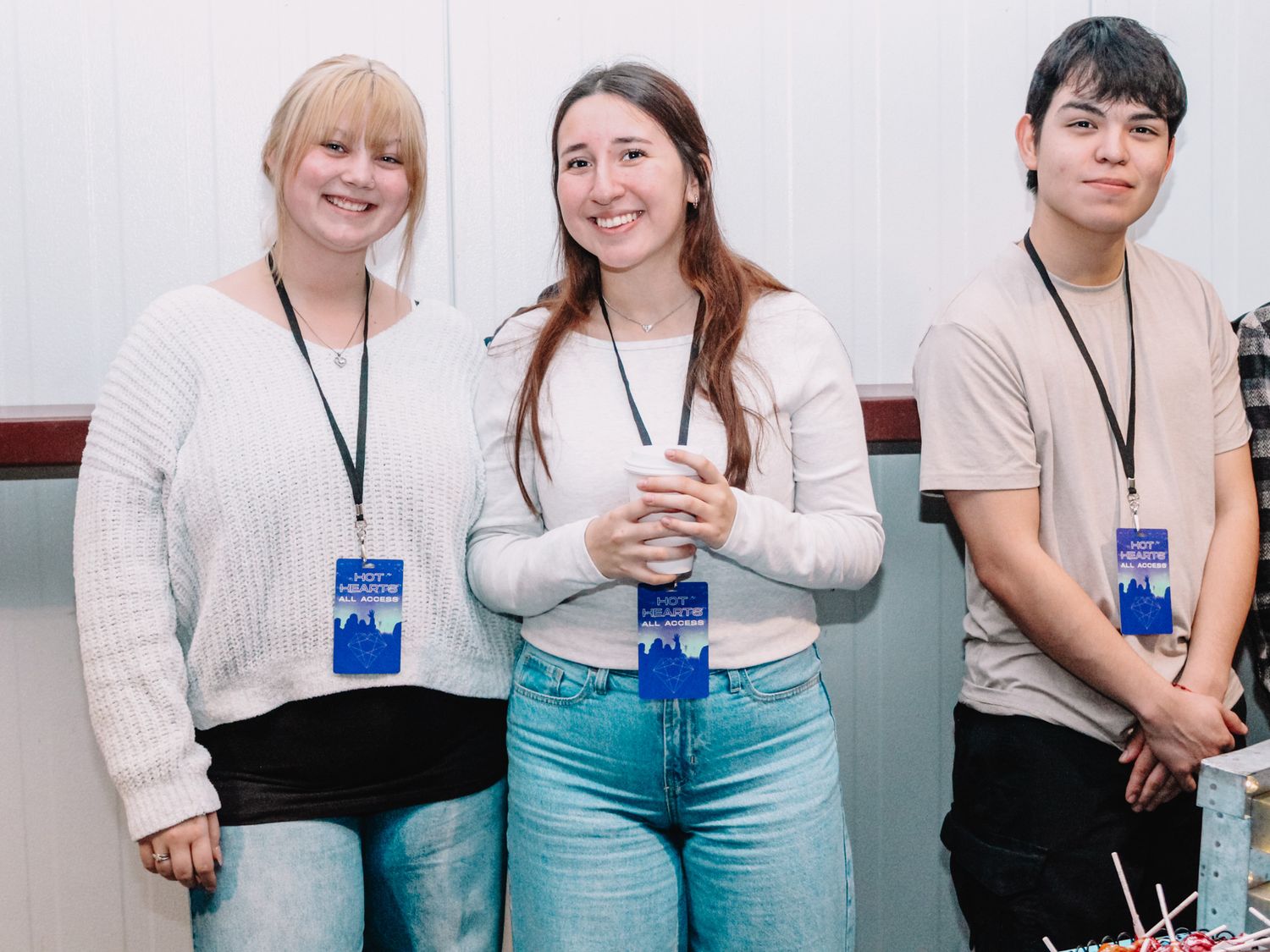 Three smiling students wearing lanyards and name badges pose together at an indoor event or conference.