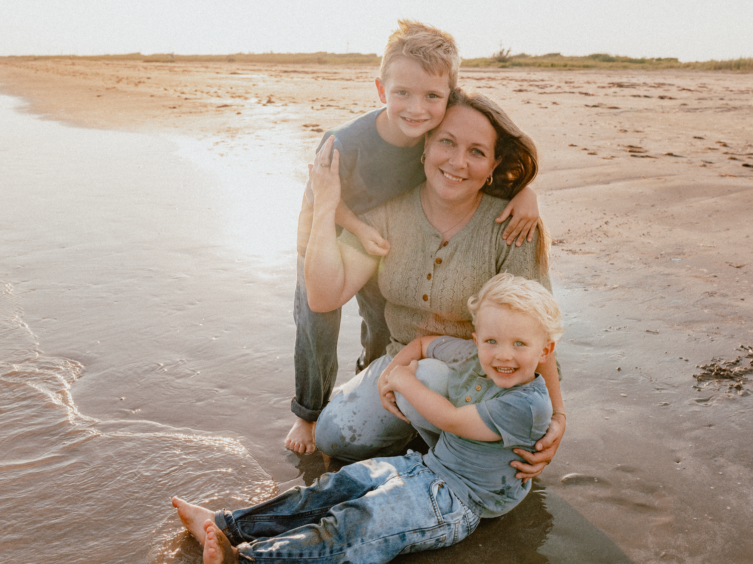 A mother smiles with her two young children on a sunny beach, sitting in shallow water with golden light behind them.