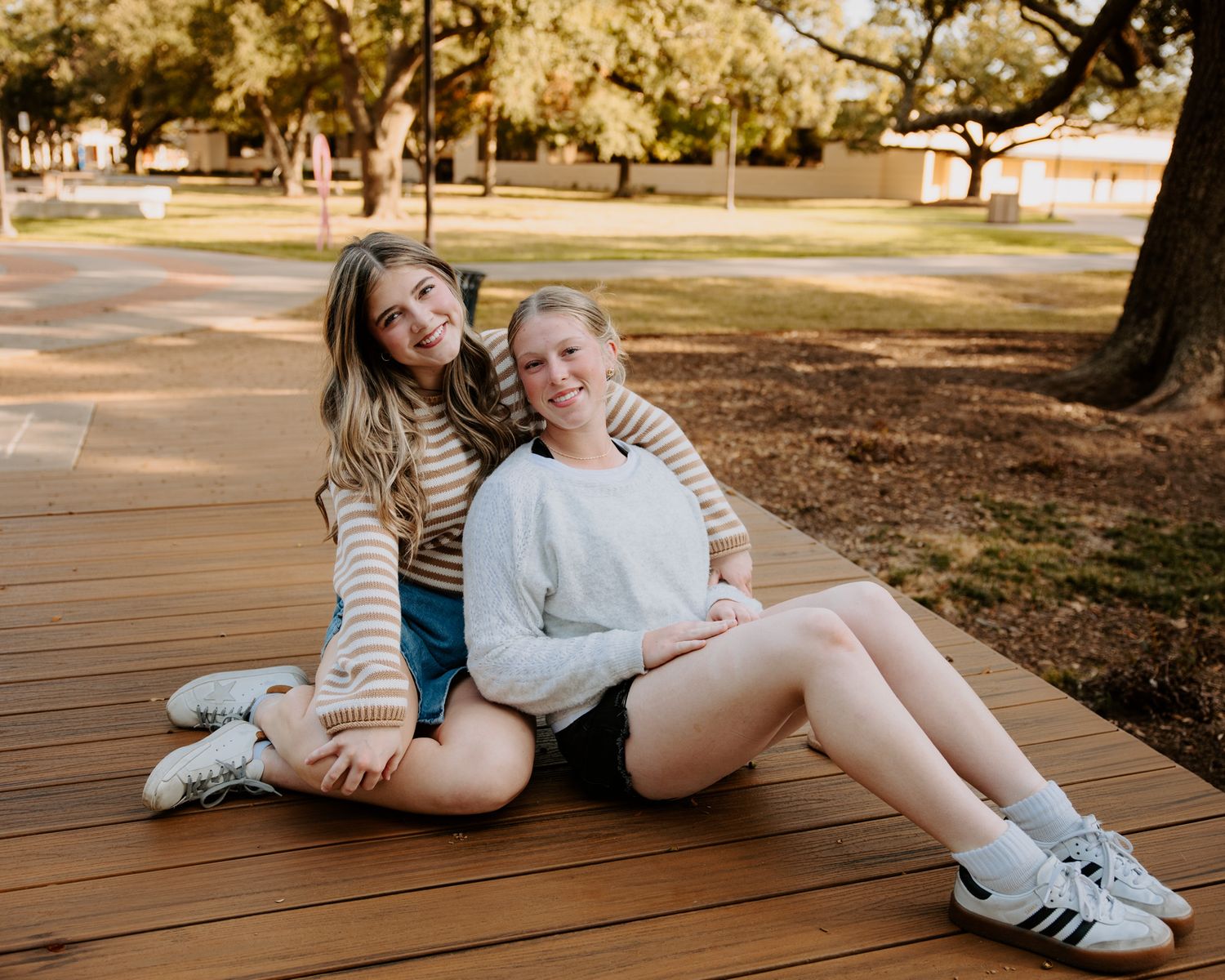 Two smiling young women sitting together on a wooden deck outdoors in a park setting.
