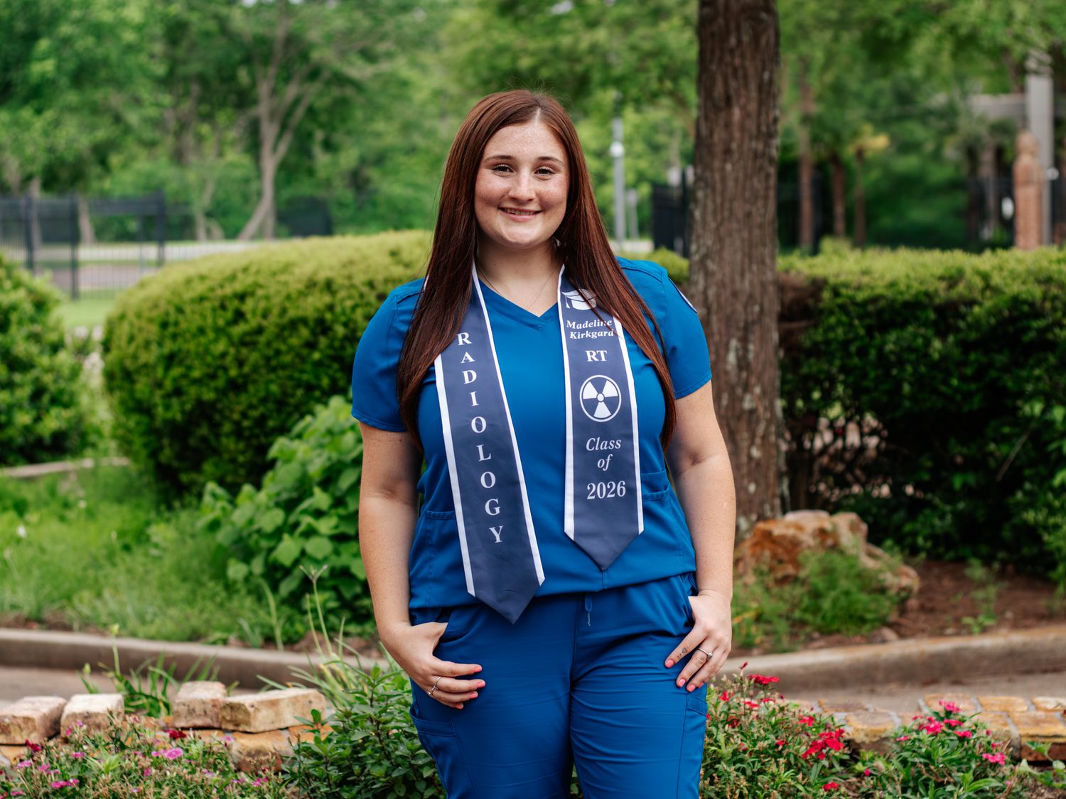 Radiology graduate smiling in blue scrubs and graduation stole, posing outdoors among green hedges and red flowers.